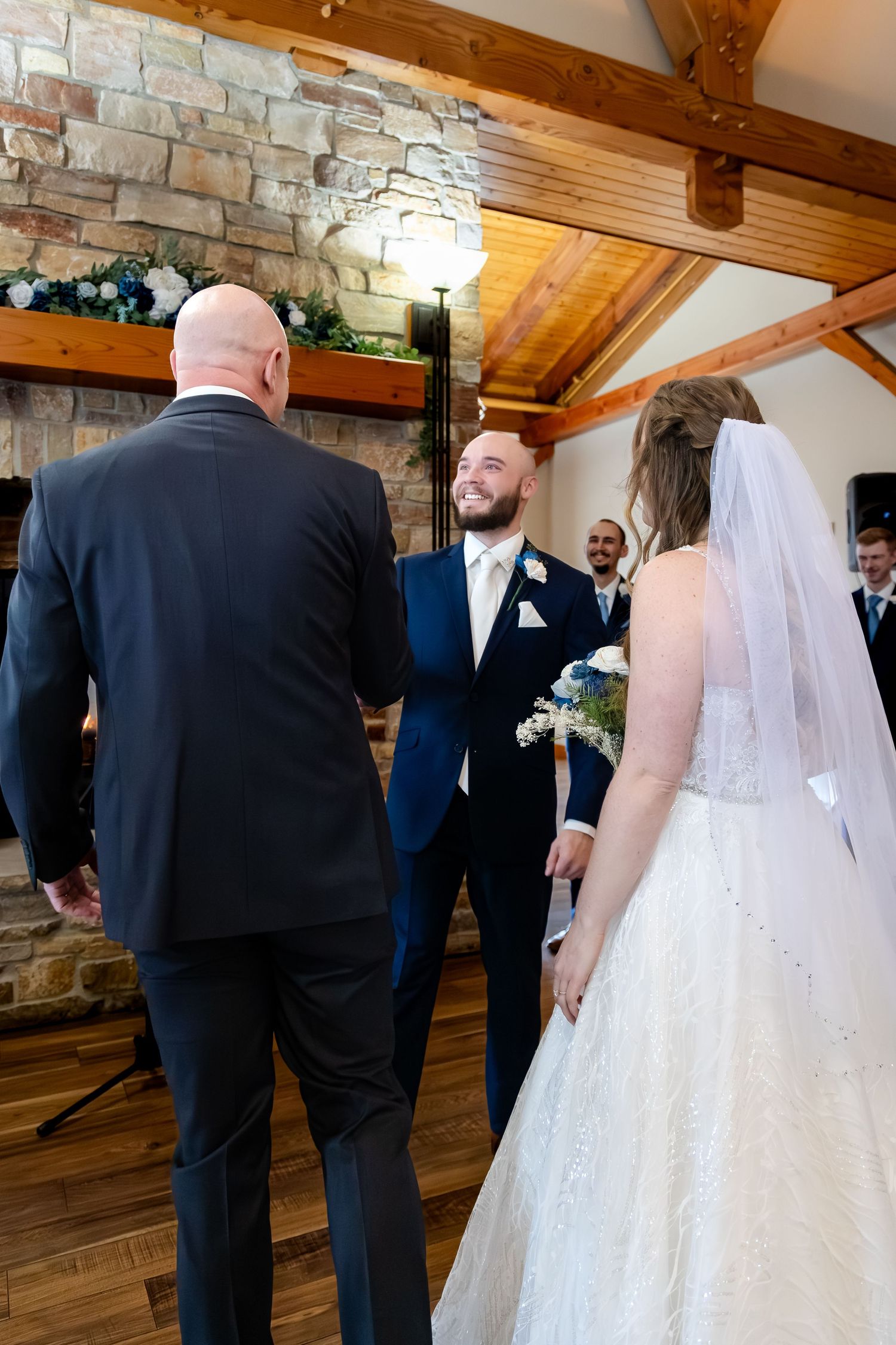 Groom in blue suit smiles at bride in white gown during indoor wedding ceremony with stone fireplace backdrop.