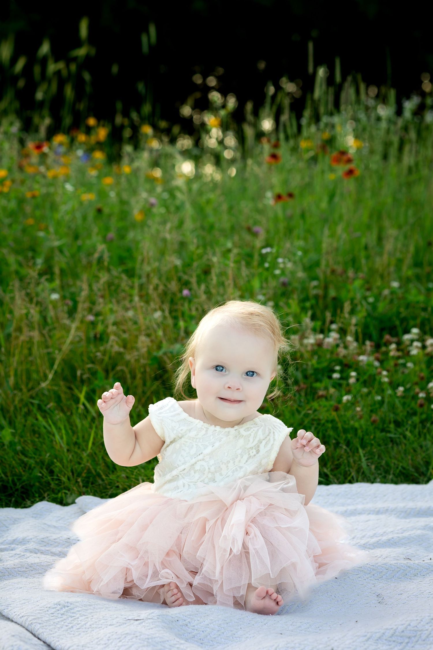 Baby in pink tulle dress sits on white blanket surrounded by wildflowers.
