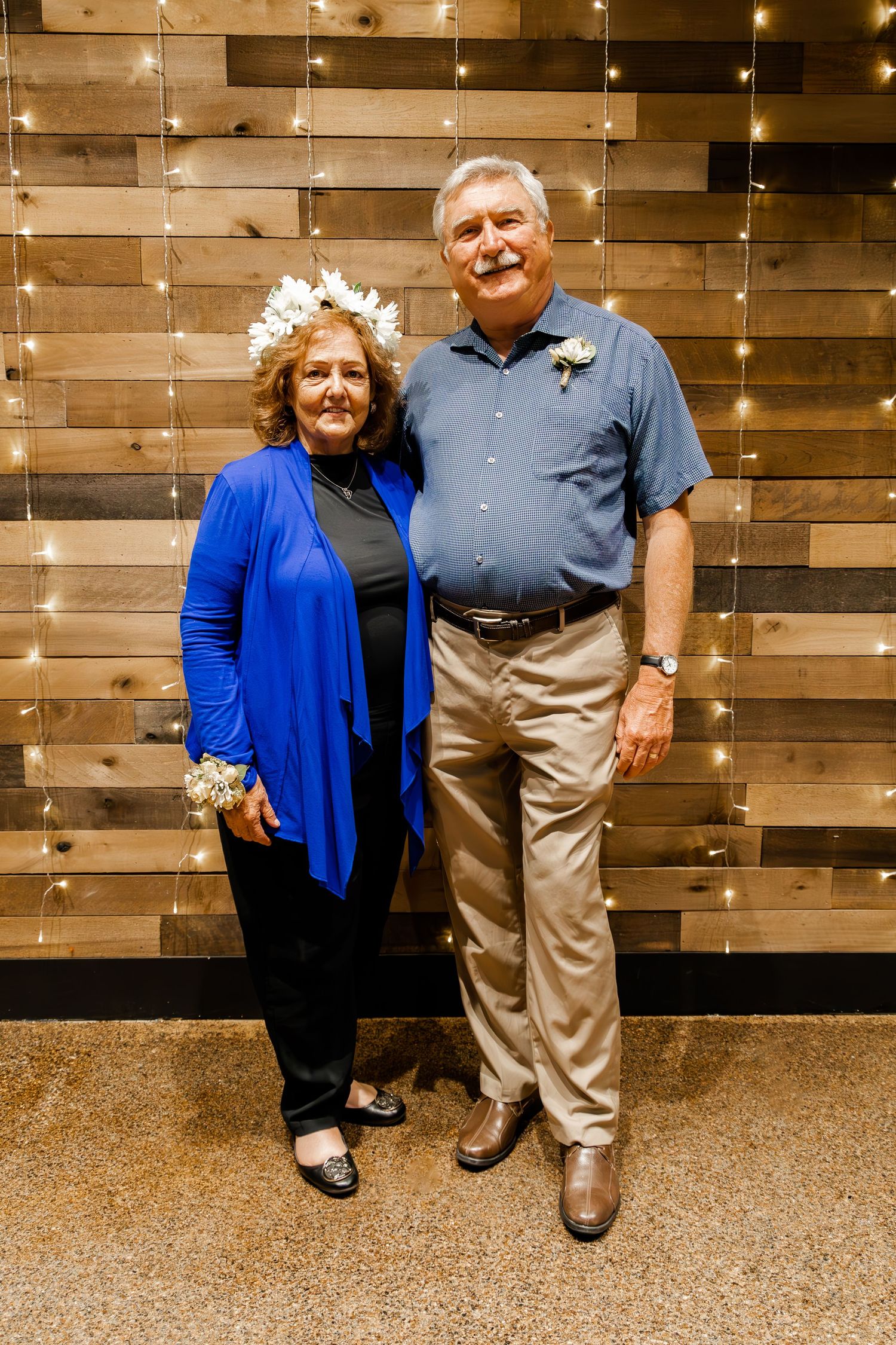 A couple poses in formal attire against a wooden wall backdrop with twinkling lights.