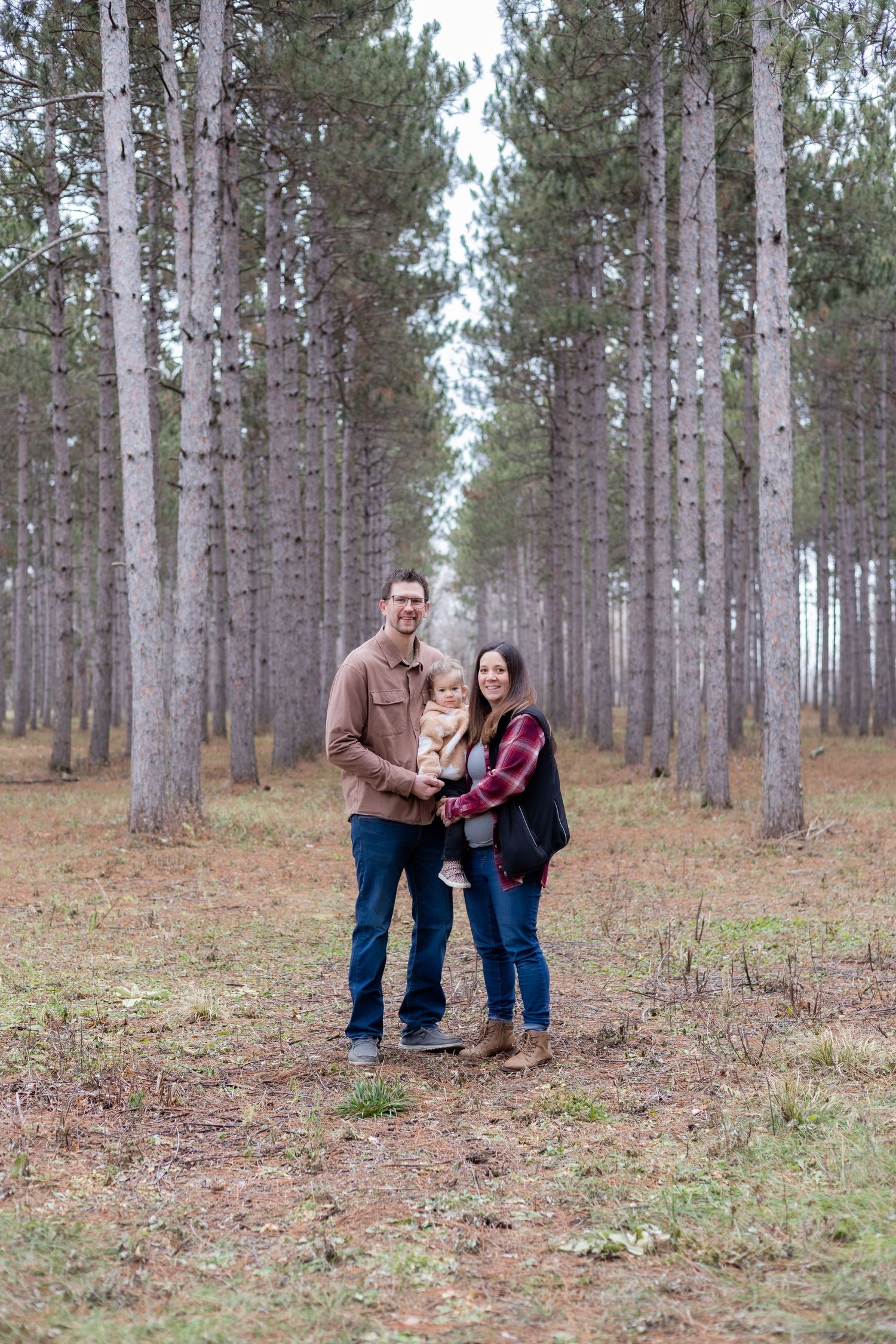 A couple stands together in a serene pine forest setting during autumn.