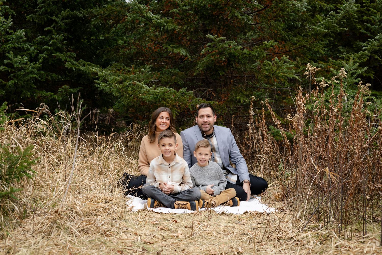A family shares candid moments together while sitting in a grassy field surrounded by evergreen trees.