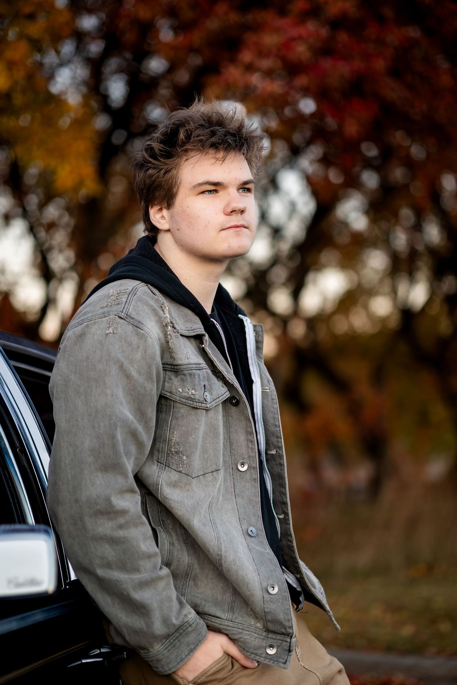 Young person in gray jacket leans against black car during autumn sunset with blurred trees in background.