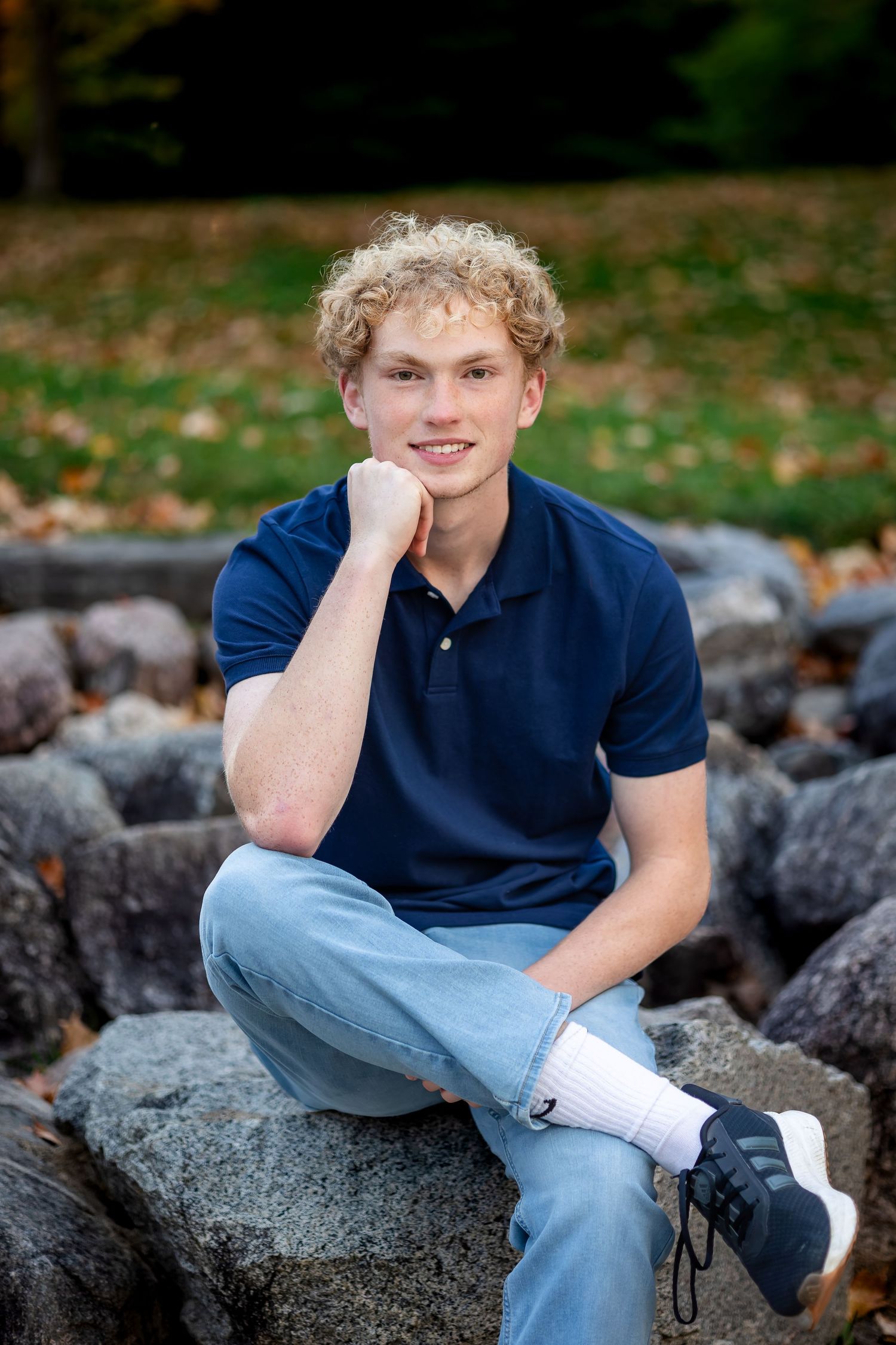 A person in a navy polo shirt and jeans sits casually on large rocks surrounded by autumn leaves.