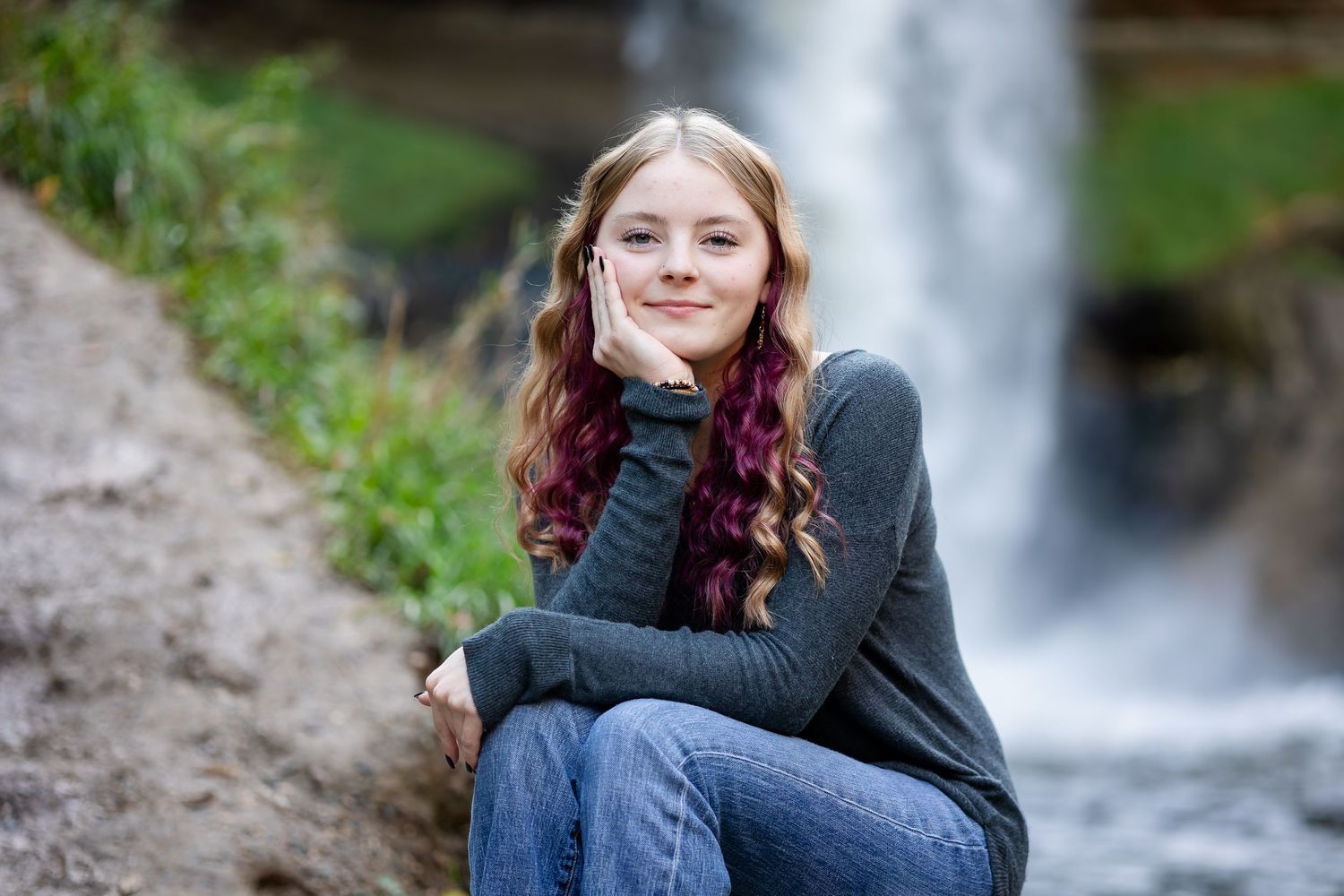 A person sits on rocks near a cascading waterfall in a forest setting wearing casual attire.