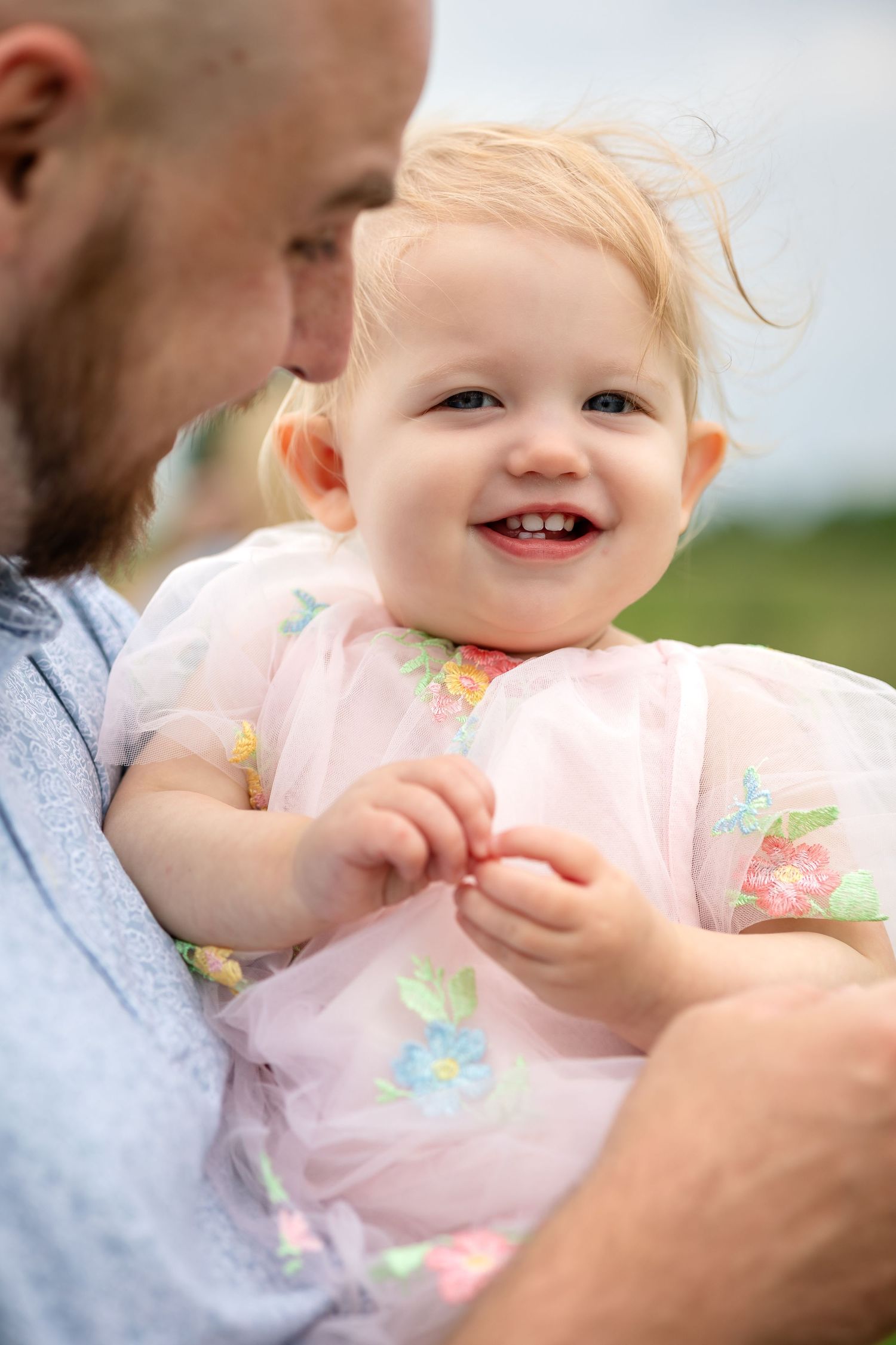 Smiling baby girl in pink floral dress being held by bearded father outdoors.
