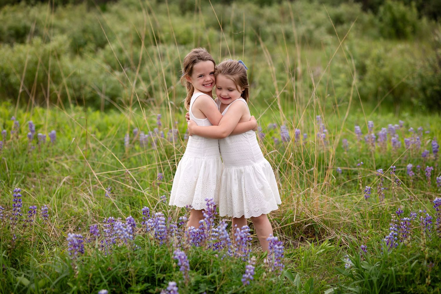 Two young girls in white dresses hugging in a field of purple lupine flowers.