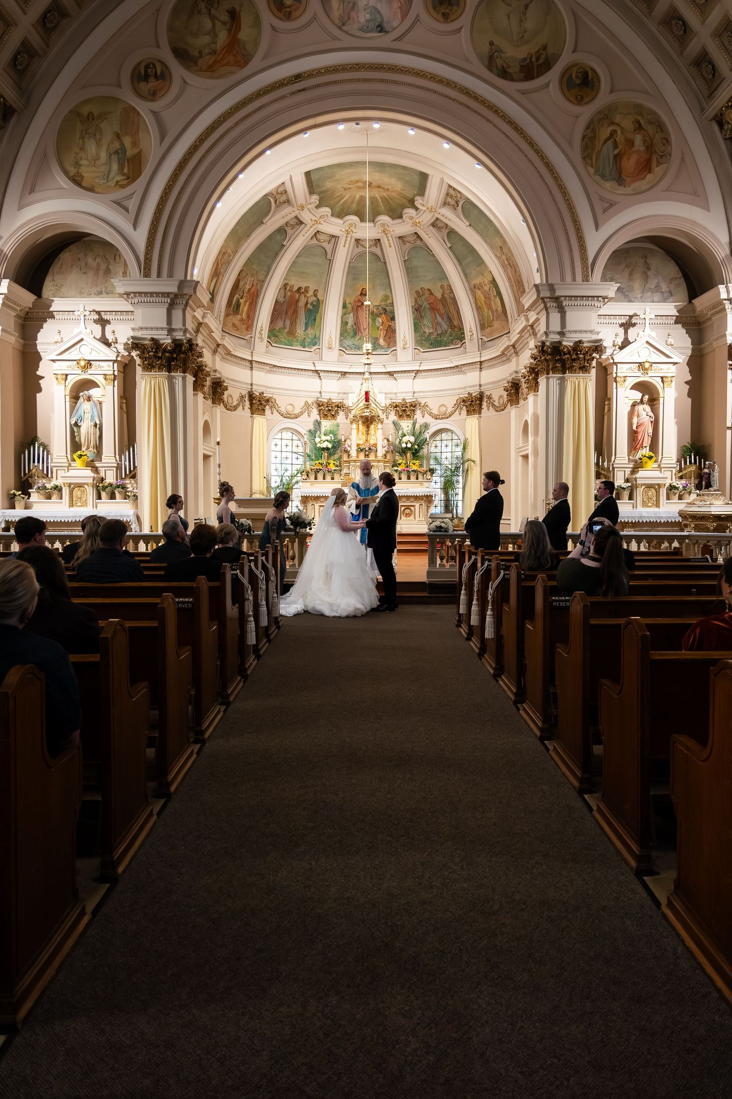 Wedding ceremony in ornate church with frescoed dome, couple at altar flanked by guests in wooden pews.