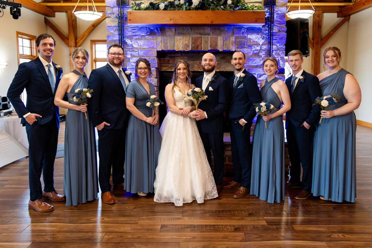 Wedding party poses in front of a stone fireplace, bride and groom centered, bridesmaids in blue gowns, groomsmen in navy suits.