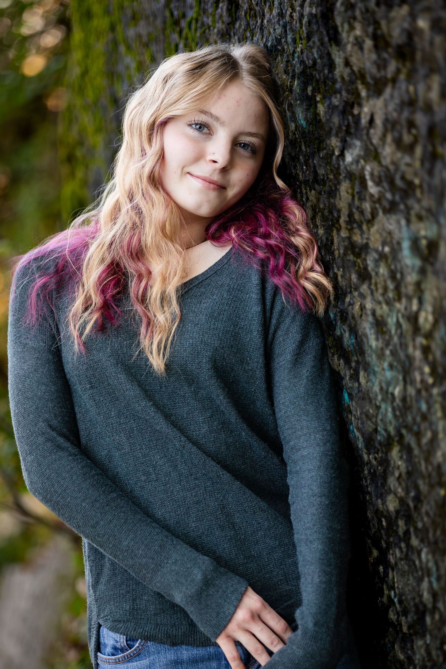 Portrait of someone with blonde and pink hair wearing a gray sweater leaning against a tree trunk in natural lighting.
