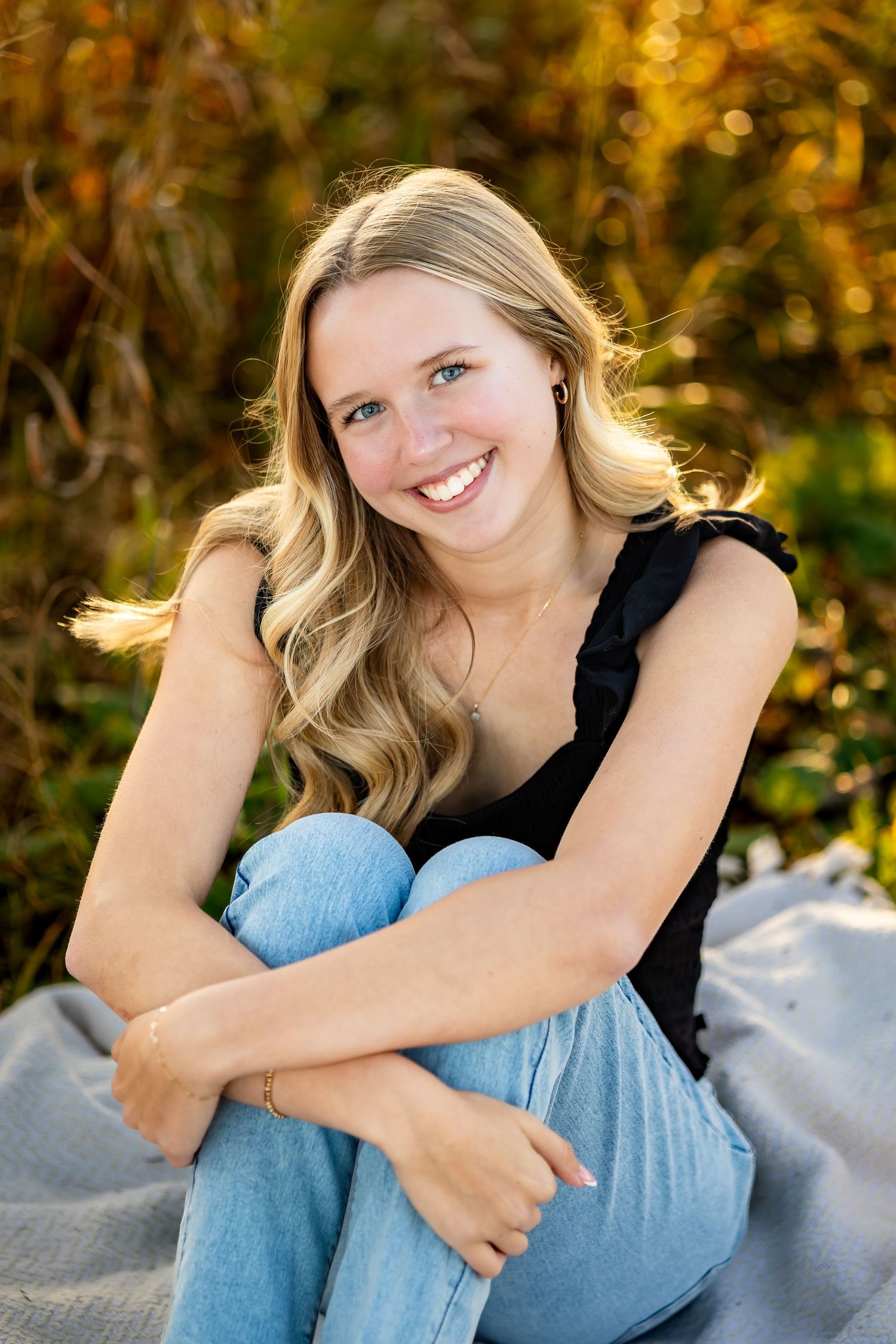 A portrait of someone in a black top and jeans sitting on a blanket with golden autumn foliage in the background.