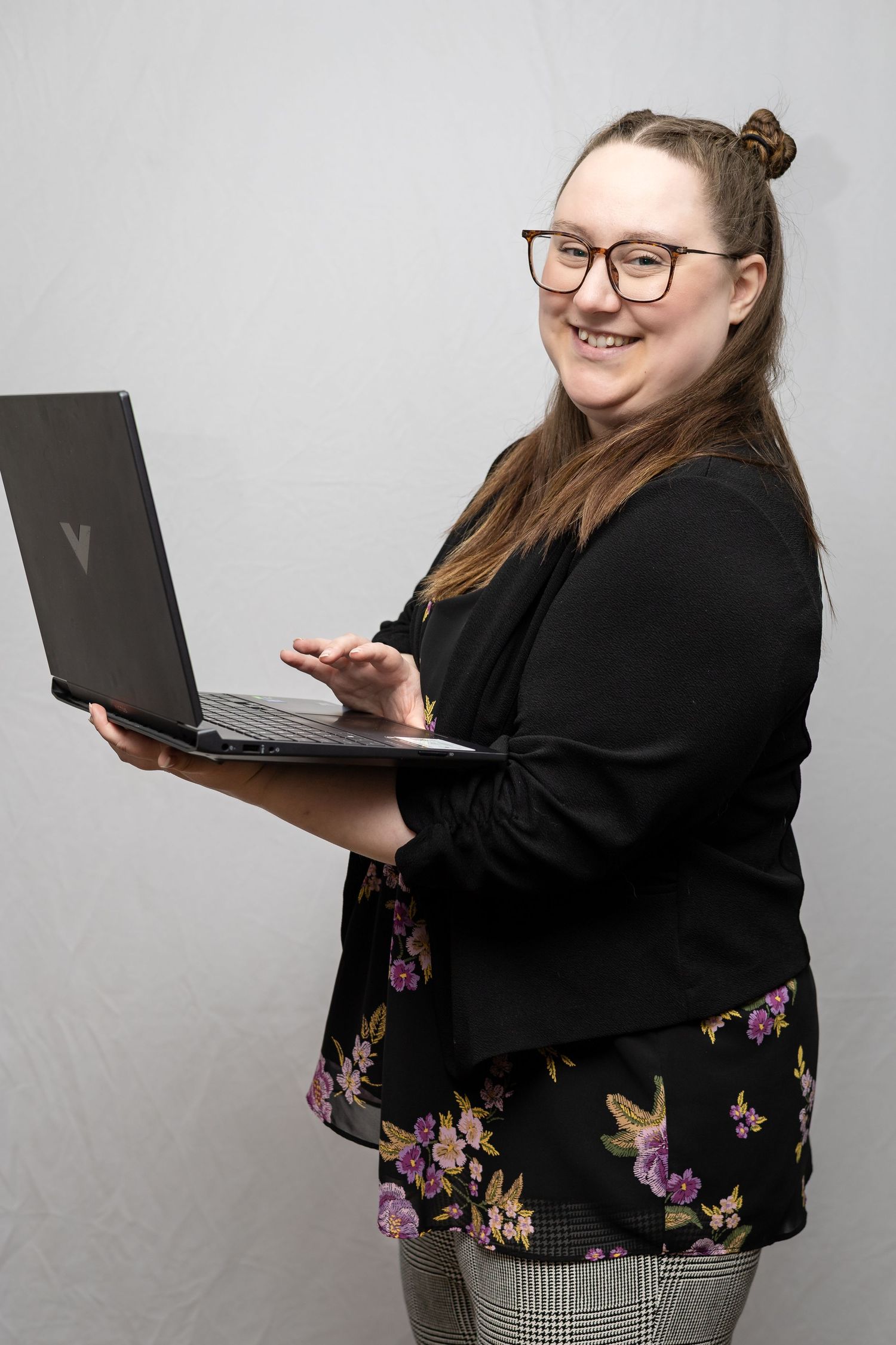 Professional in a black blazer and floral top holds a laptop while posing against a gray studio background.
