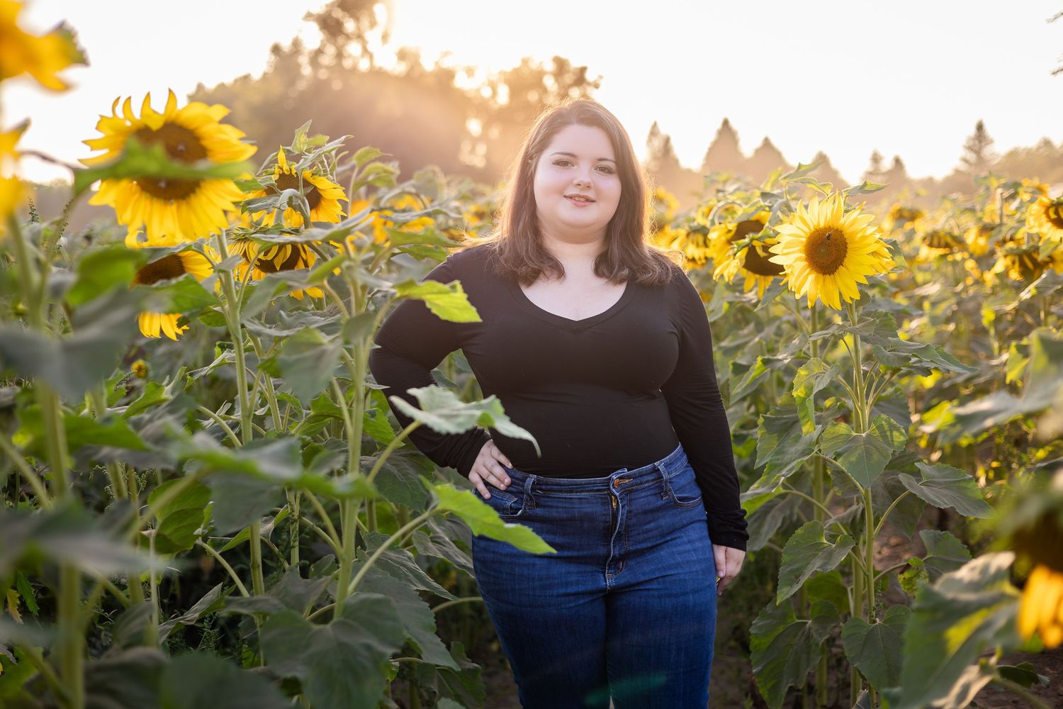 Woman in black top stands among tall sunflowers during sunset photo session.