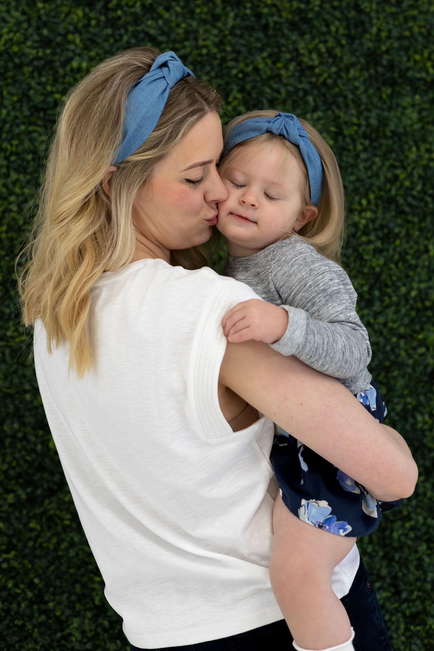 A tender moment between parent and child wearing matching blue headbands against a green hedge backdrop.