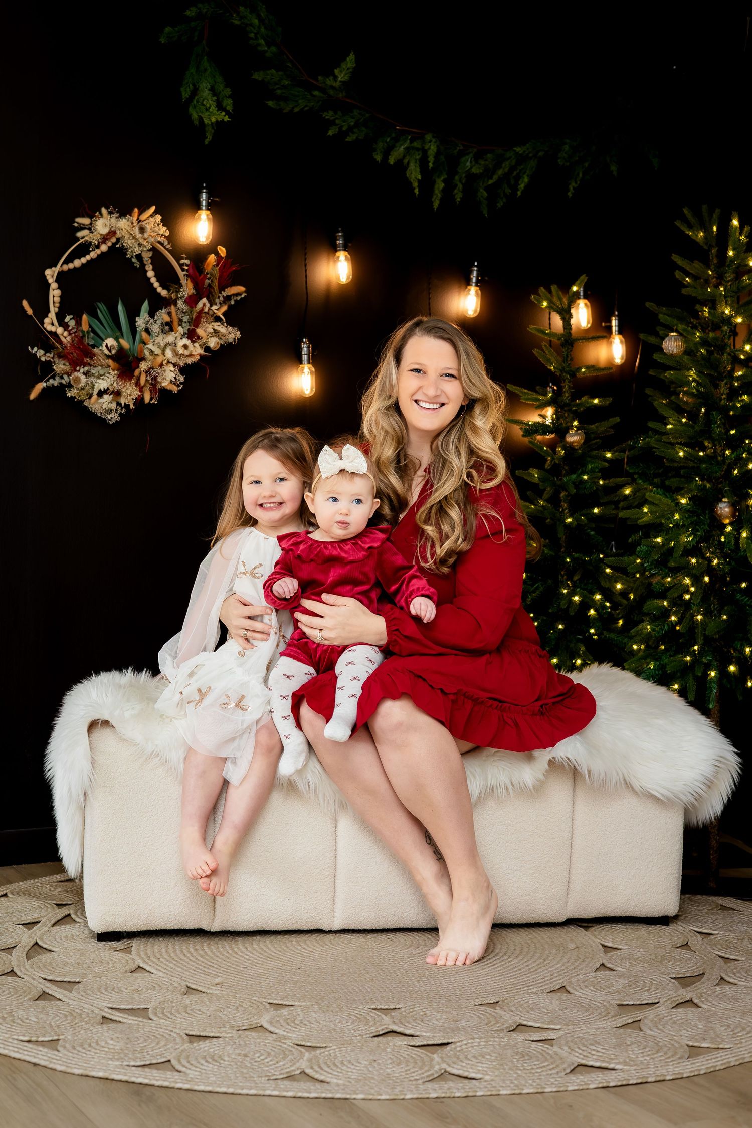 A smiling mother in a red dress sits on a white sofa with her two young daughters in a festive Christmas setting.