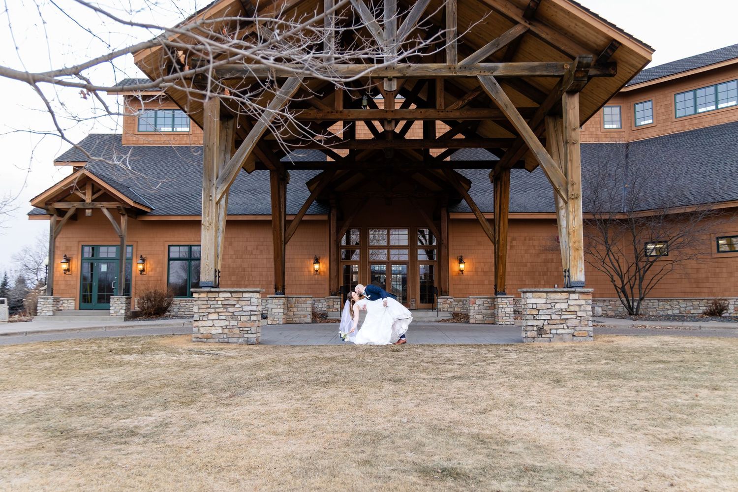 Bride and groom embrace in front of a rustic lodge wedding venue with timber architecture on a winter day.