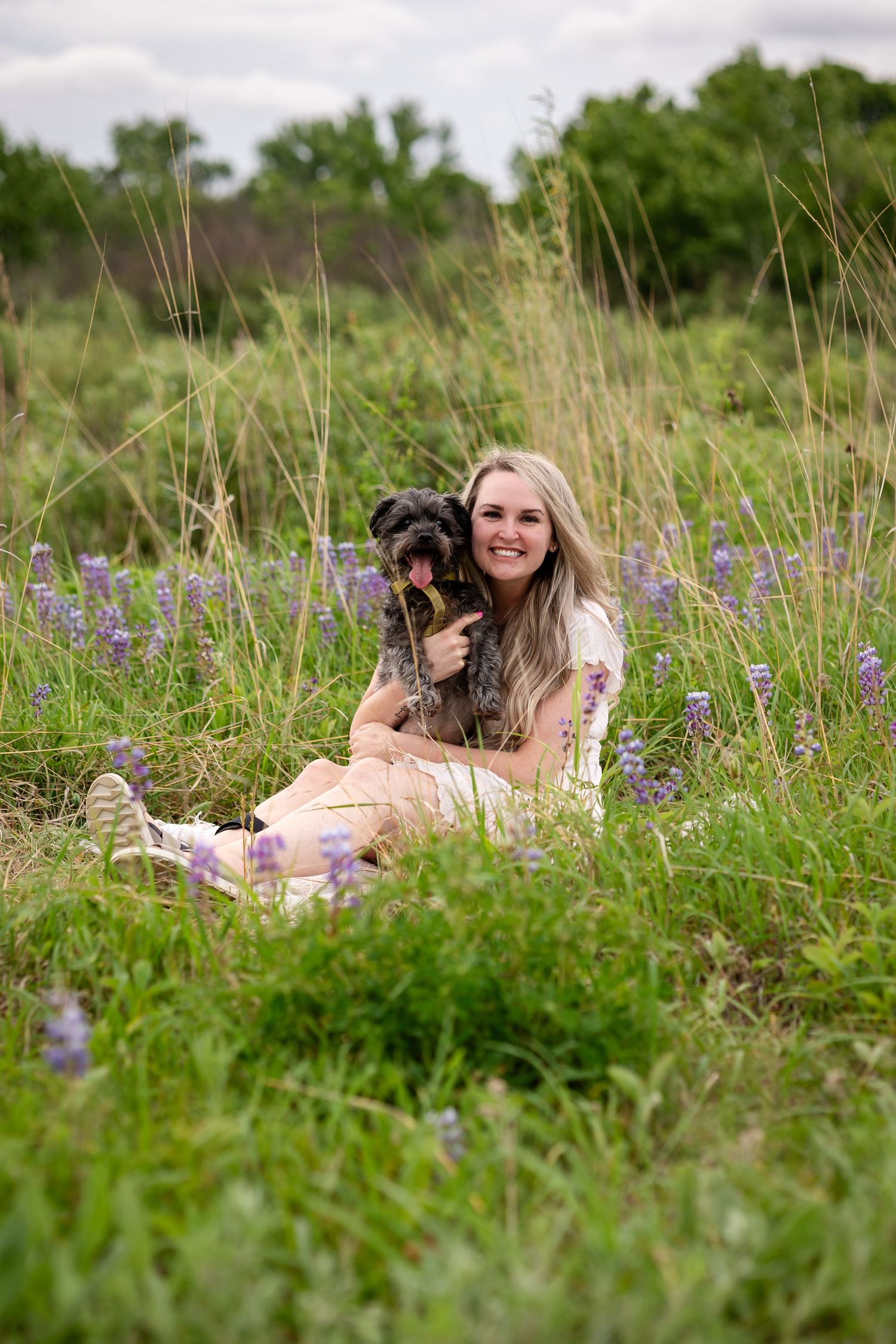 Young woman with long blonde hair sitting in a wildflower field, smiling while hugging a small black dog.
