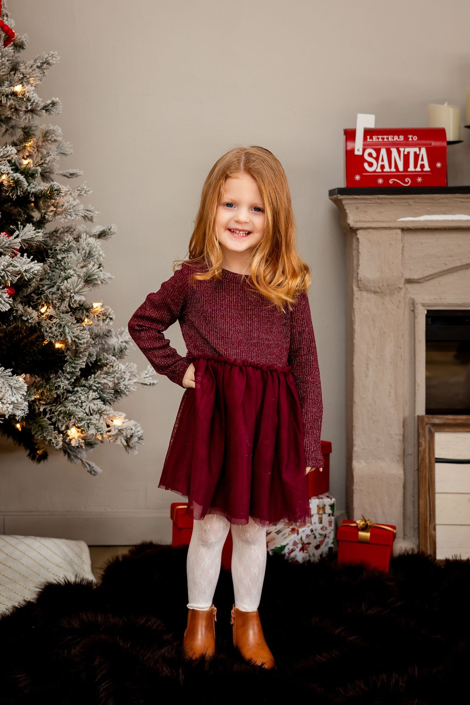 Young girl in burgundy dress poses by Christmas tree with Santa letterbox and fireplace.
