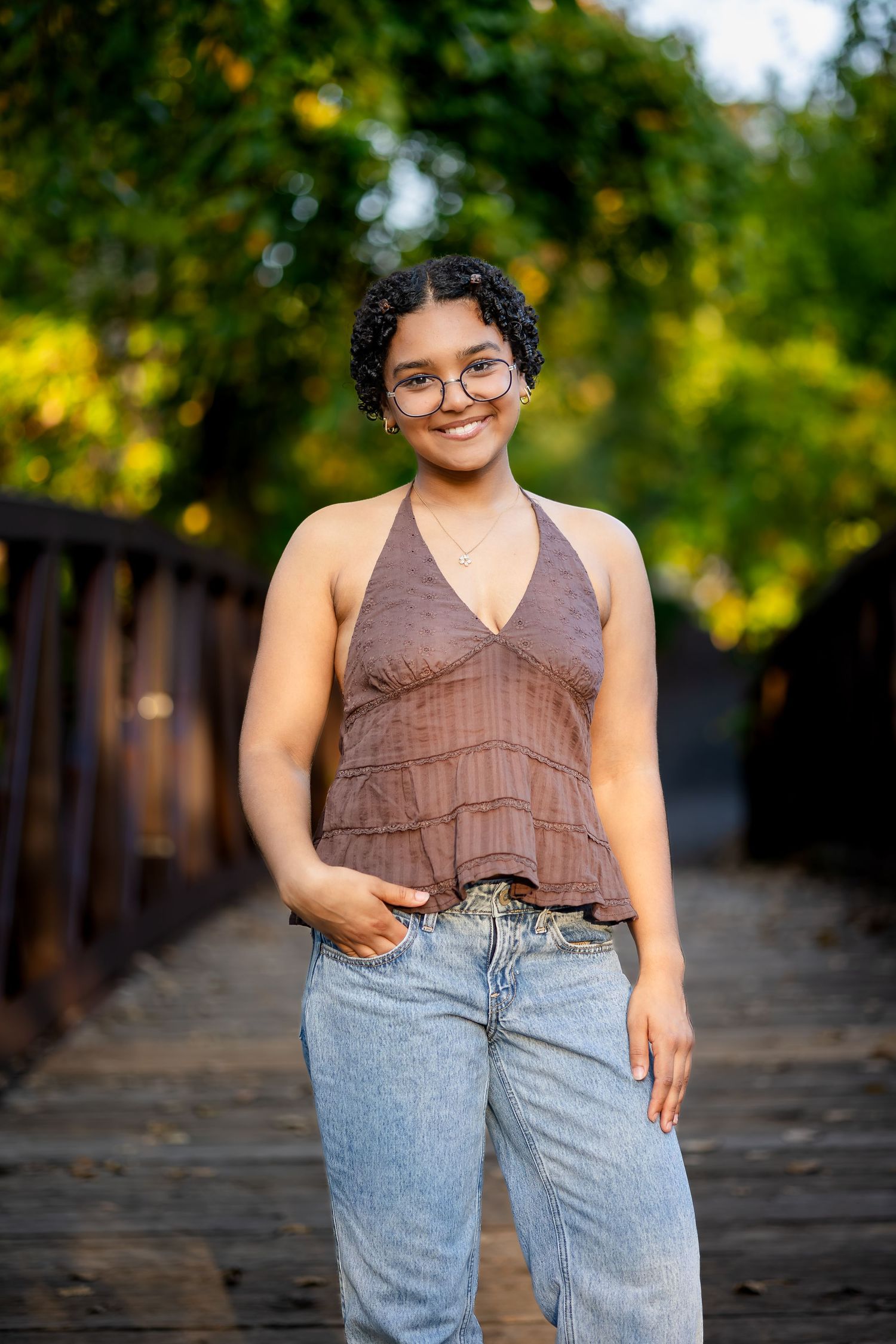 A person in a brown sleeveless top and light wash jeans poses outdoors on a wooden boardwalk with trees in background.