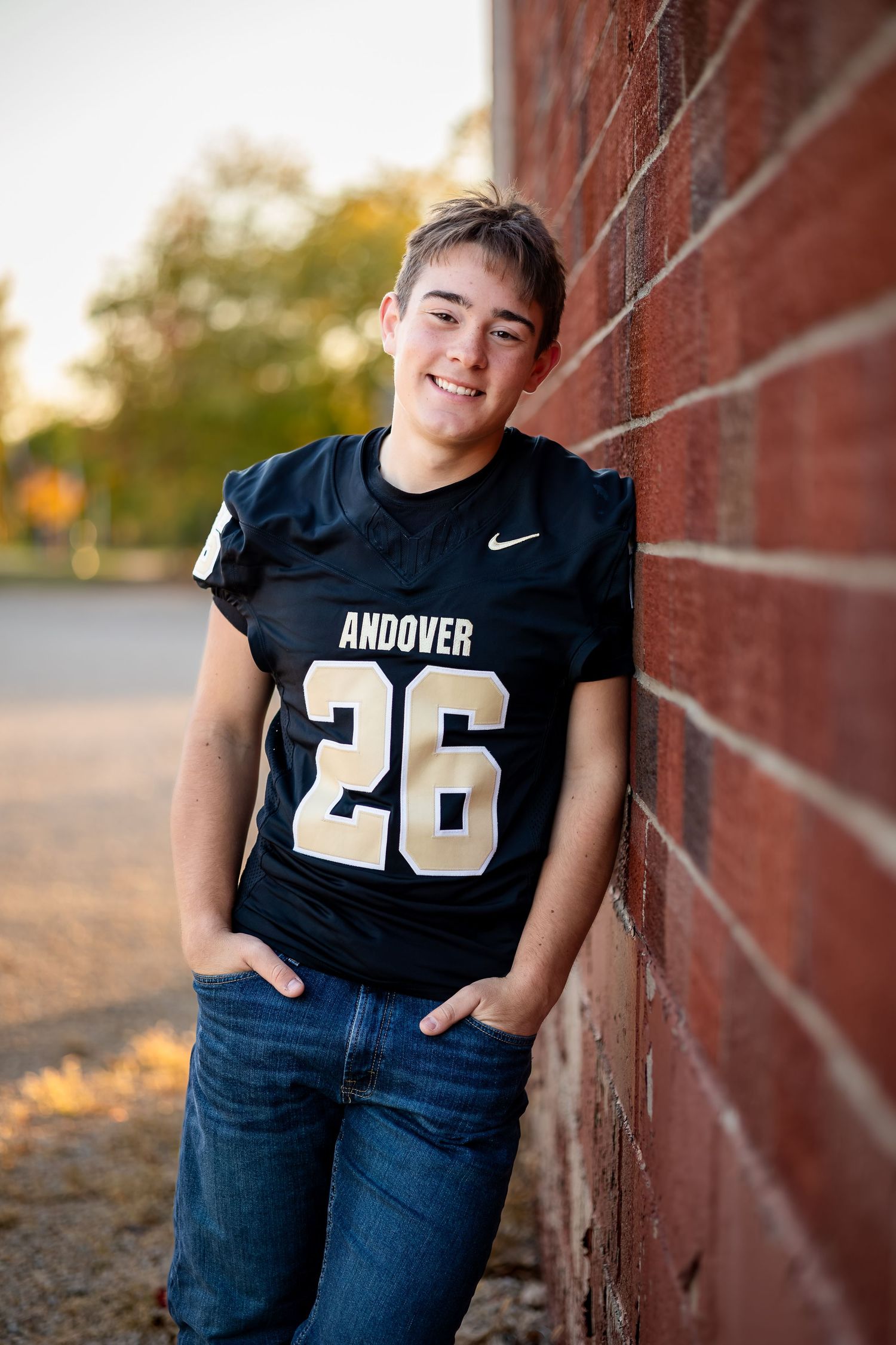 Student athlete in navy football jersey leans against brick wall during outdoor portrait session at sunset.
