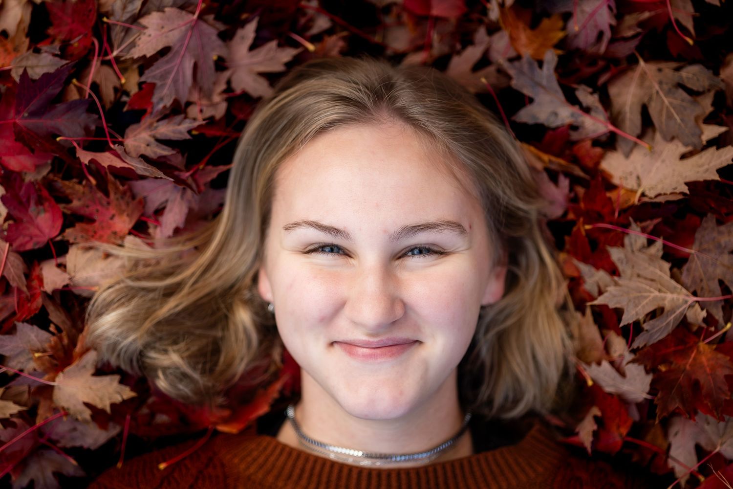 Person lying on ground surrounded by vibrant red and brown autumn leaves in outdoor setting.