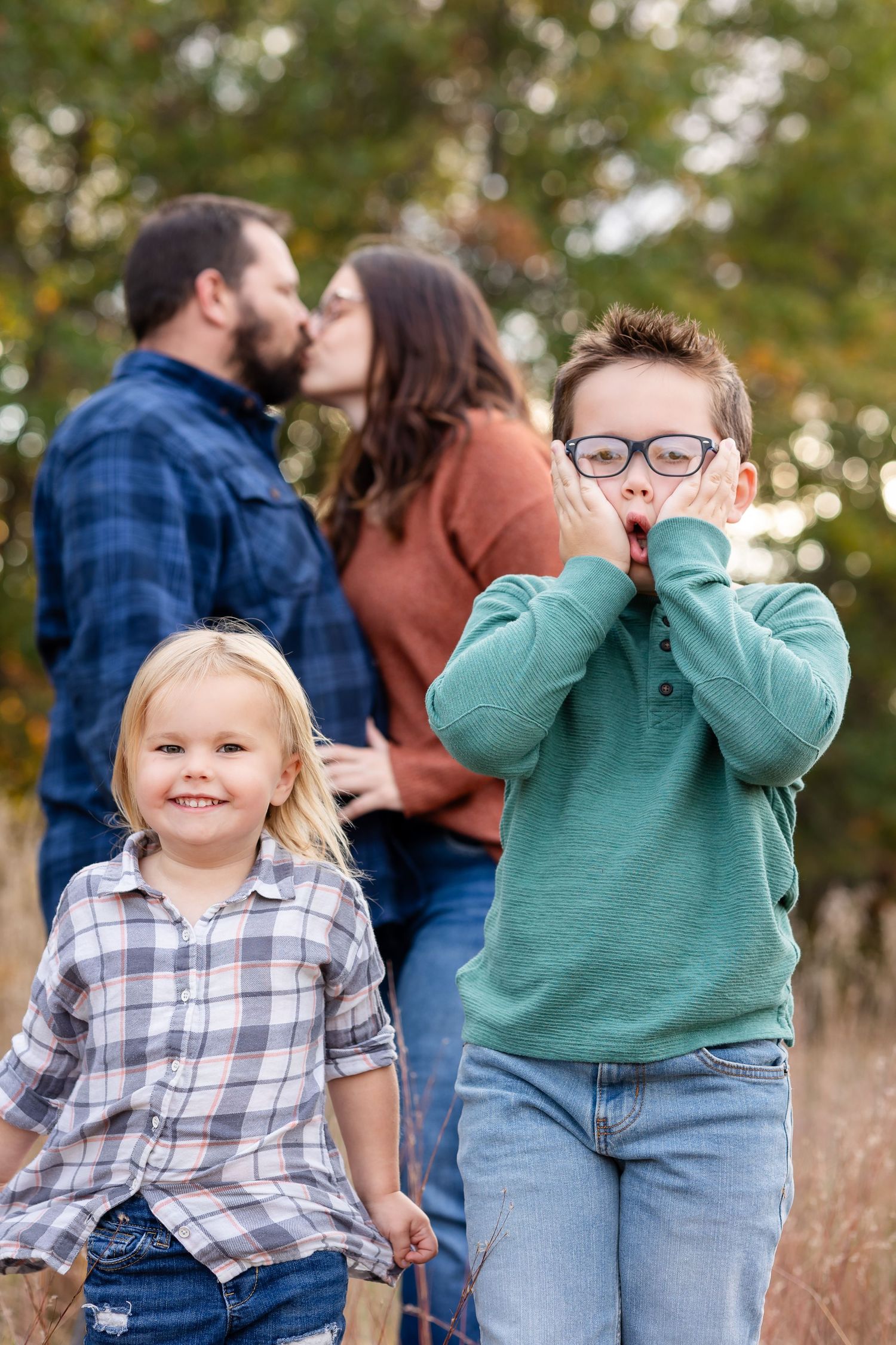 A playful family photo outdoors with children making silly faces while parents share a kiss in background.
