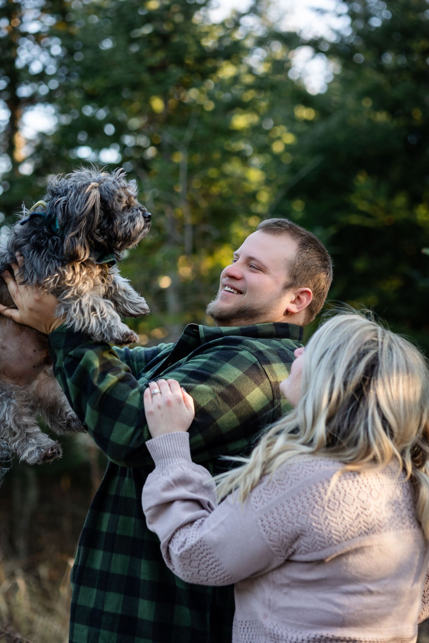 A playful outdoor photoshoot featuring a couple and their scruffy black dog among trees at sunset.