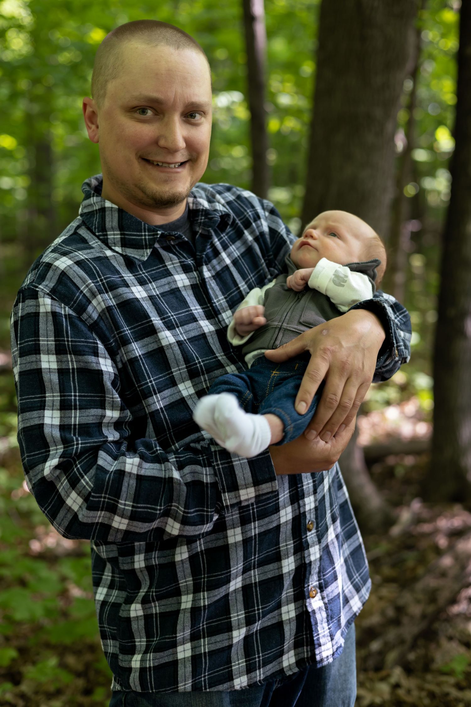 Parent in plaid shirt holding and playing with infant outdoors in wooded setting.