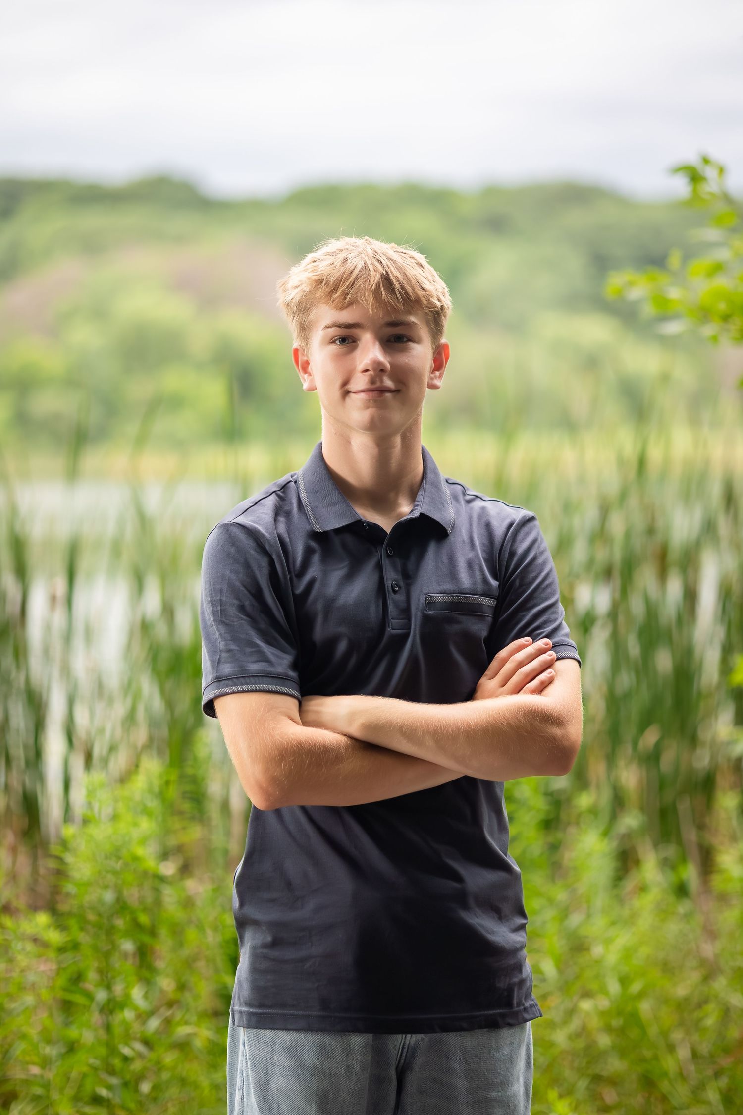 Young person in dark polo shirt standing confidently with arms crossed near lake and tall grass.