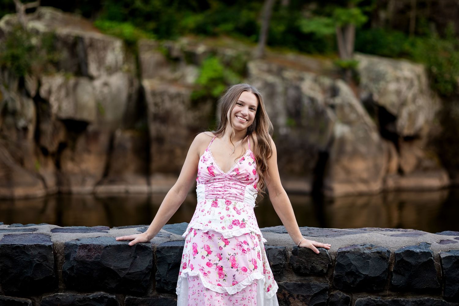Woman in floral pink dress poses elegantly against dramatic rock cliff backdrop.