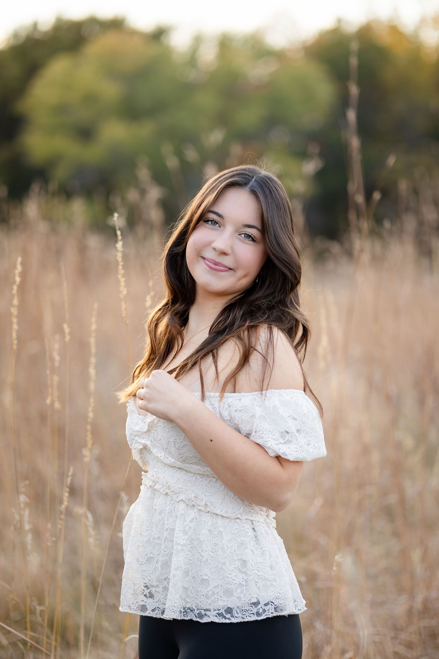Natural light portrait in white lace top among tall grass at sunset.