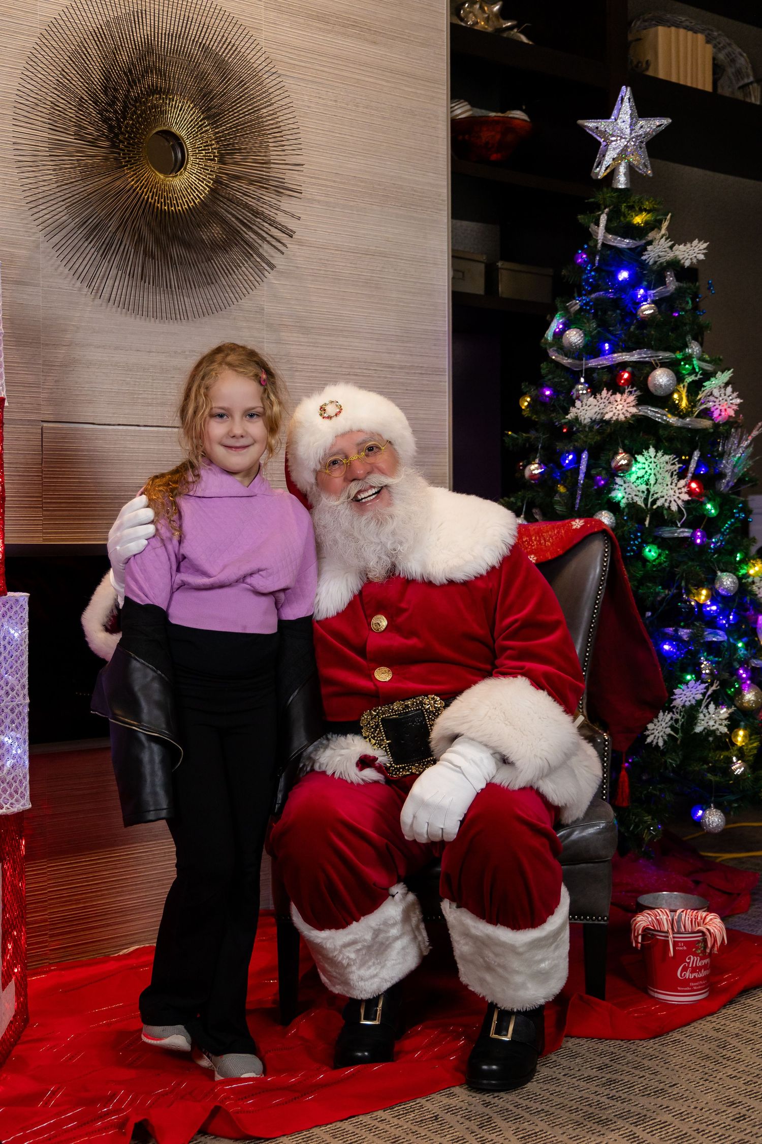 A festive Christmas scene with Santa in his traditional red suit seated by a decorated tree with colorful lights.