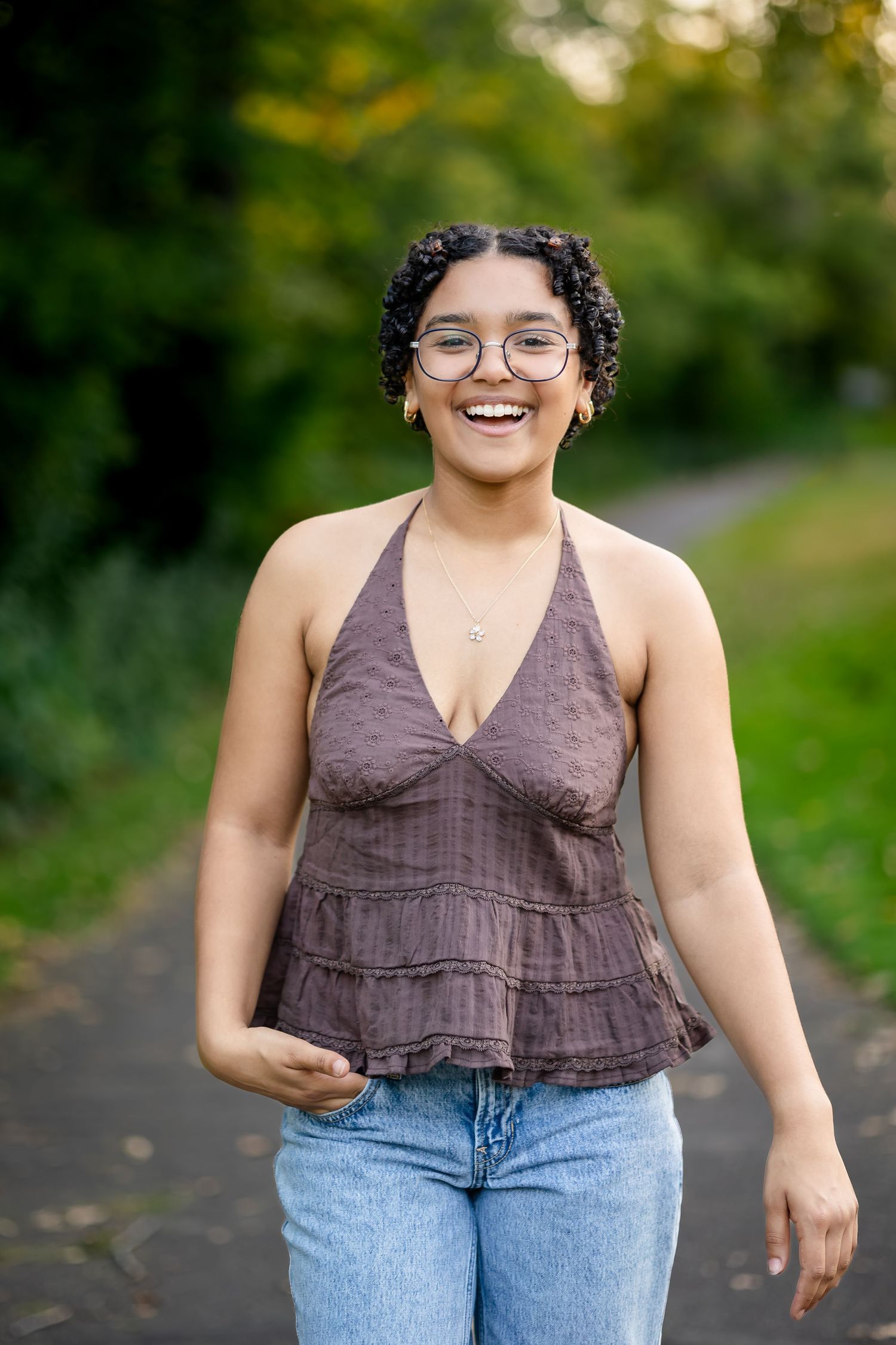 Joyful person in brown halter top and jeans walks along outdoor path with lush greenery in background.