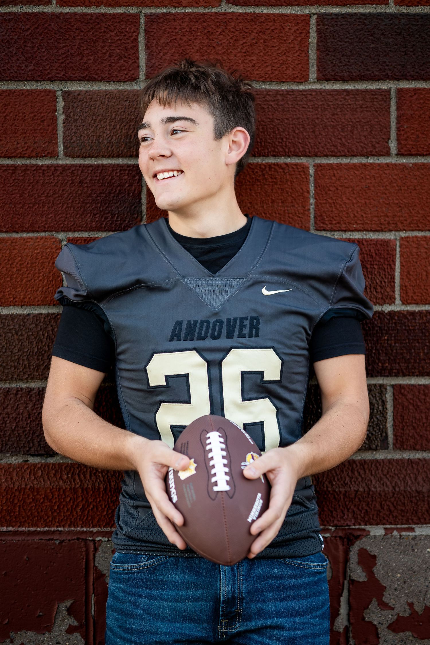 High school football player in navy Andover jersey number 26 poses with football against brick wall.