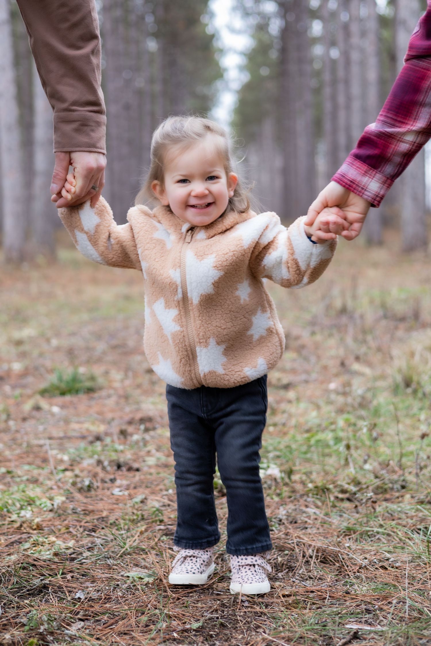 A toddler in a beige sweater holds hands with parents while walking in a forest.