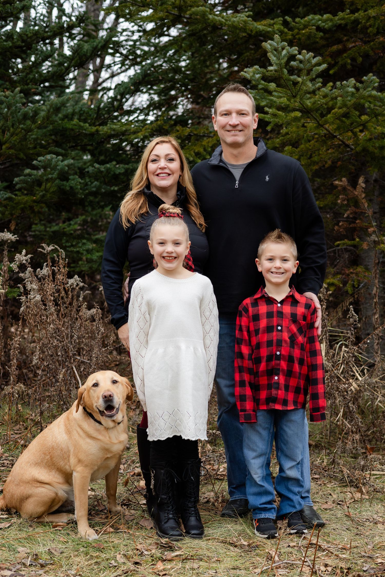 Family portrait outdoors with two children and a golden retriever dog against evergreen trees.
