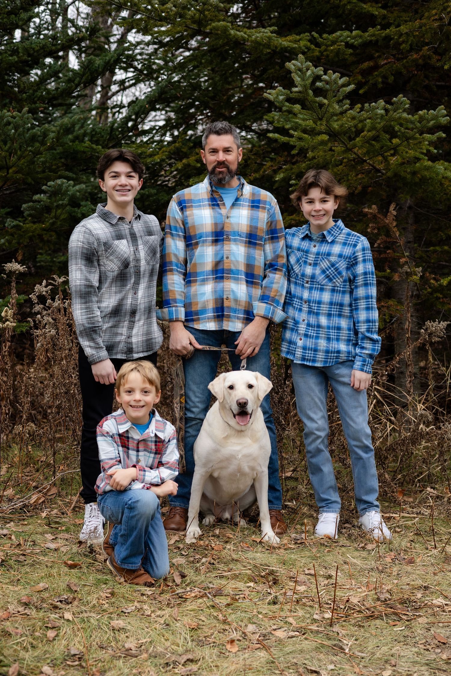 A casual outdoor family portrait with a white dog and everyone wearing coordinated blue and plaid shirts.