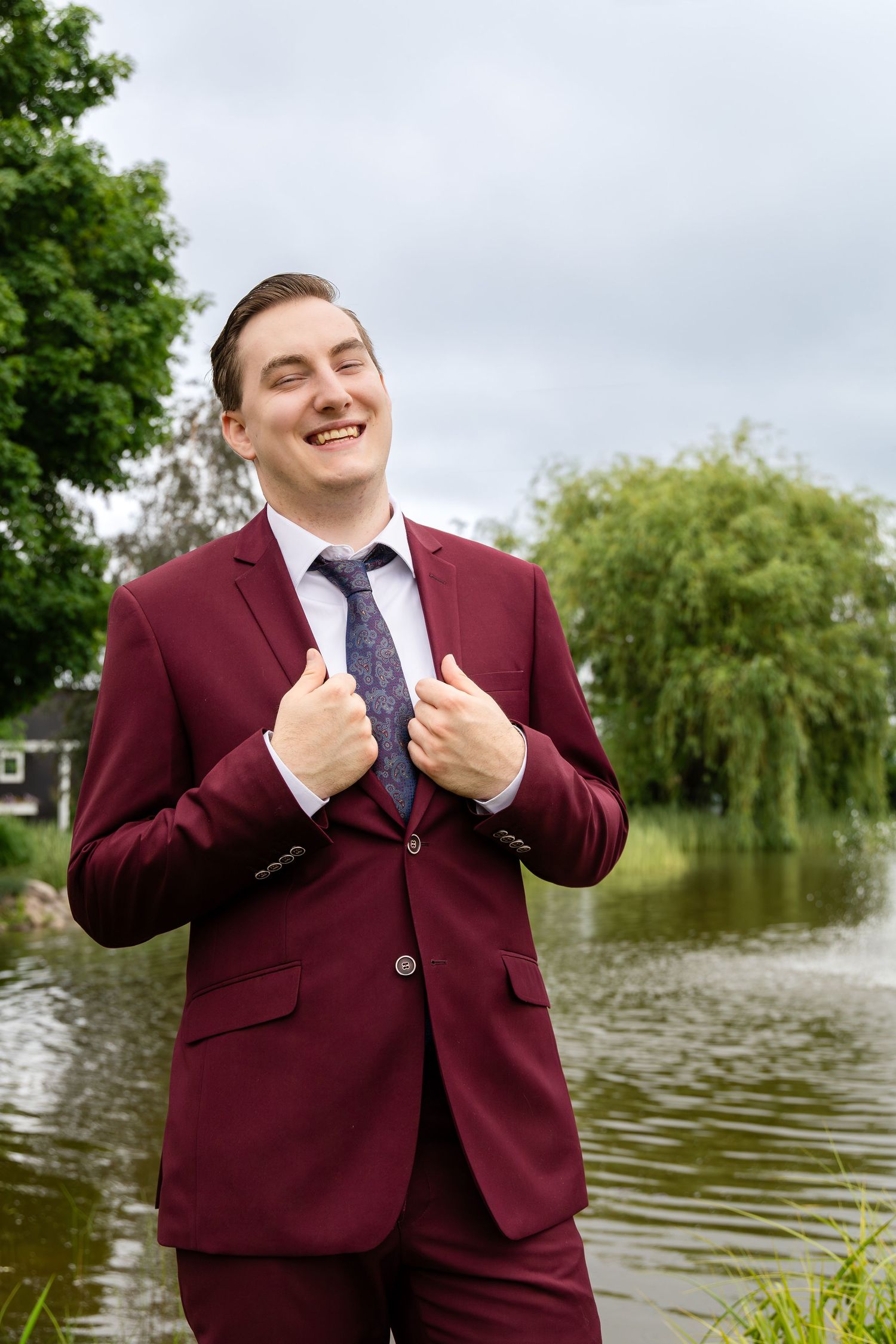 A groom in a burgundy suit poses expressively by a lake with willow trees.