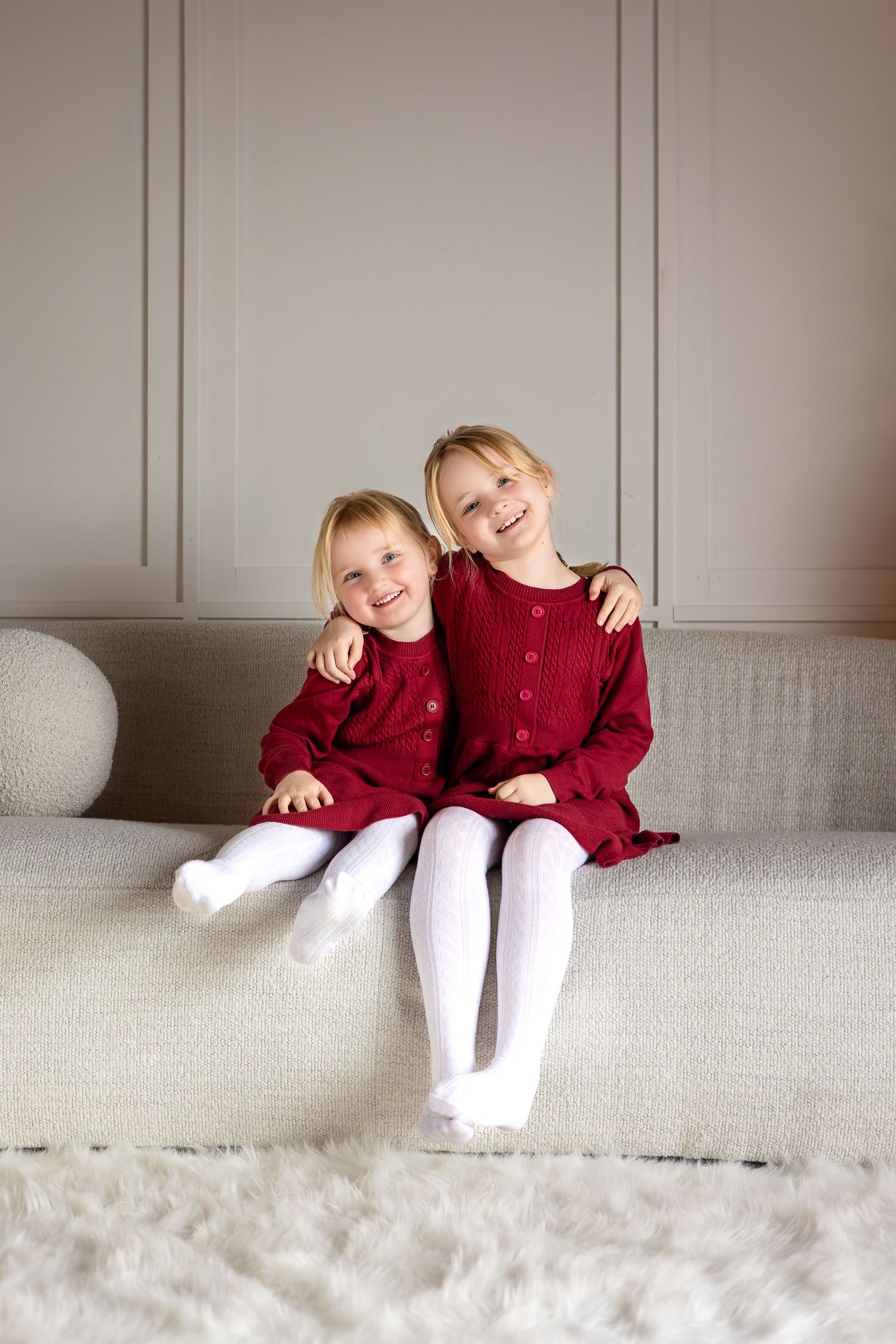 Children in matching red sweaters lounging playfully on white couch in minimalist setting.