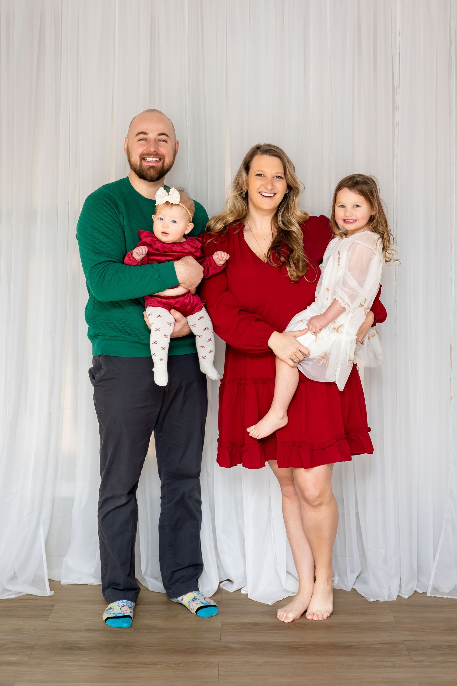 Happy family of four posing for a holiday portrait, parents holding two young daughters in red and green outfits.