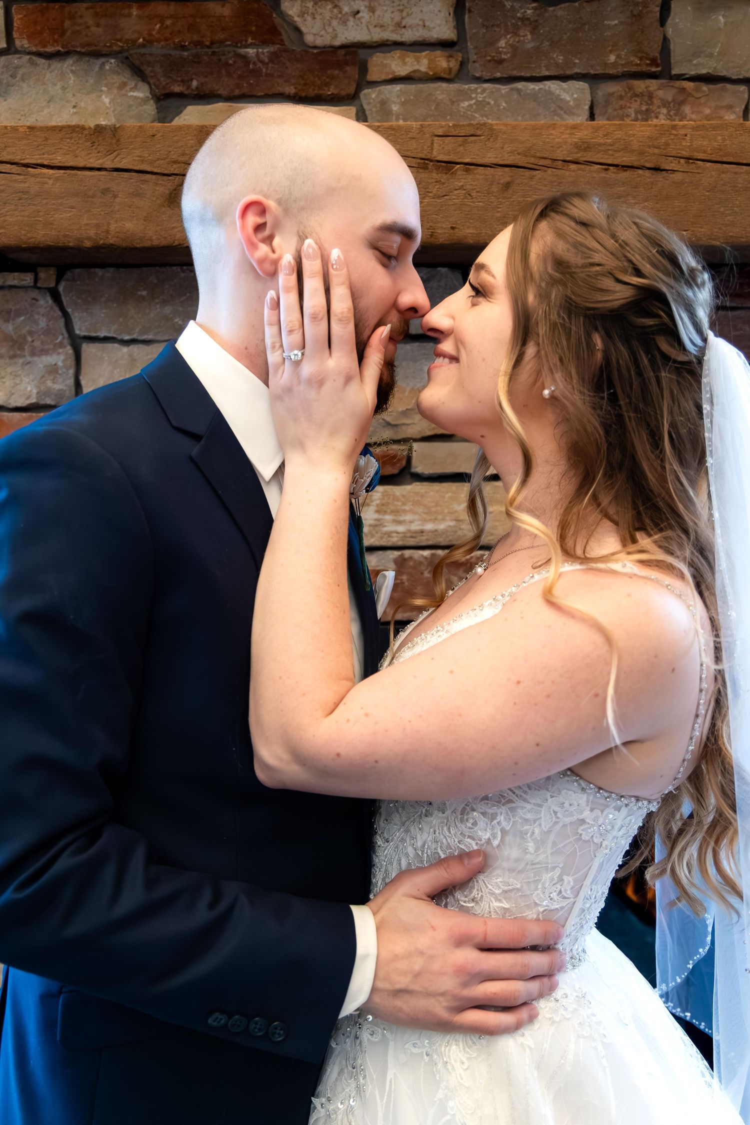 Bride and groom share a romantic kiss near a rustic stone fireplace on their wedding day.