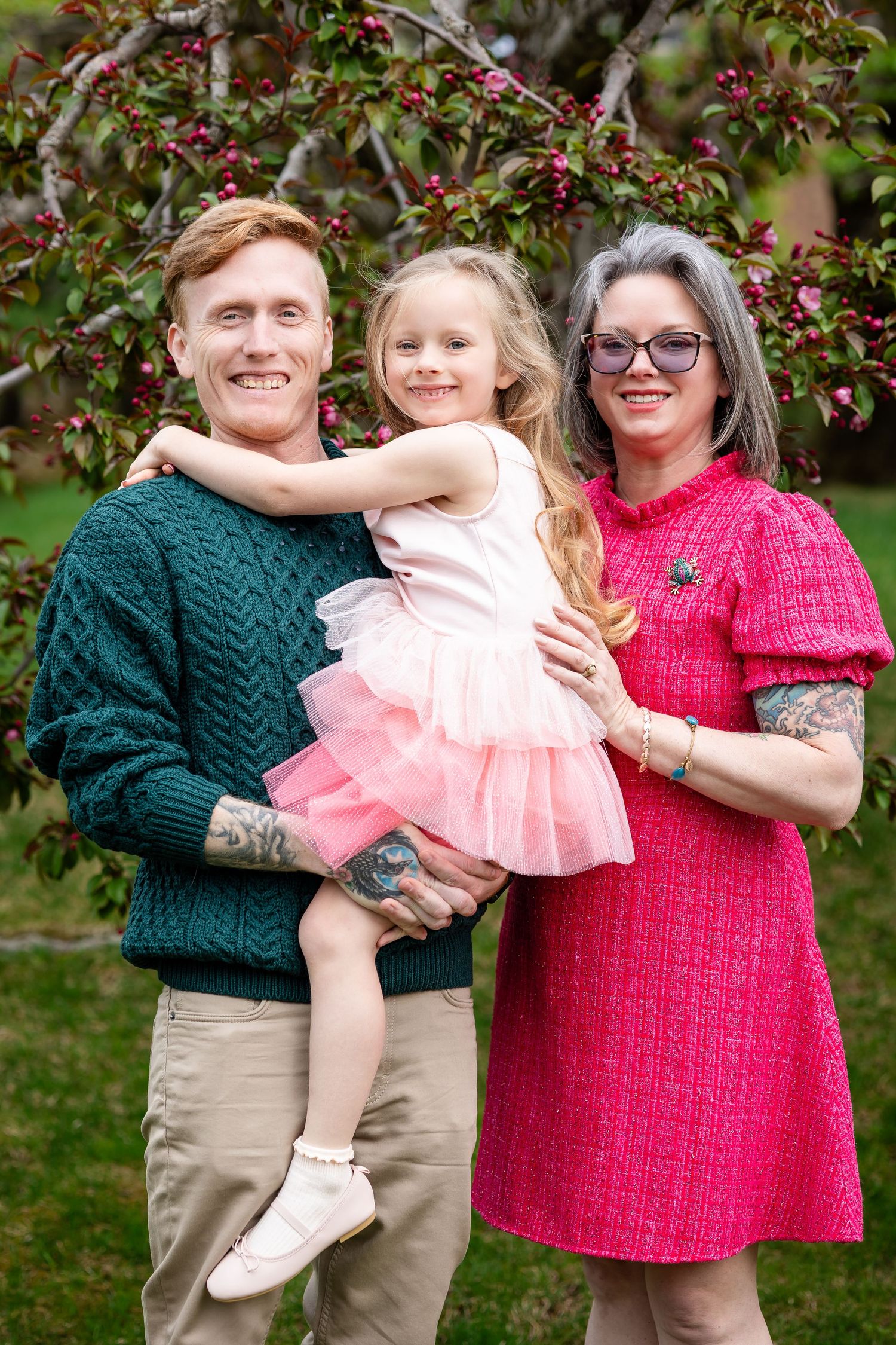 Family of three posing outdoors near flowering tree, man in green sweater and woman in pink dress holding young girl in pink tutu.