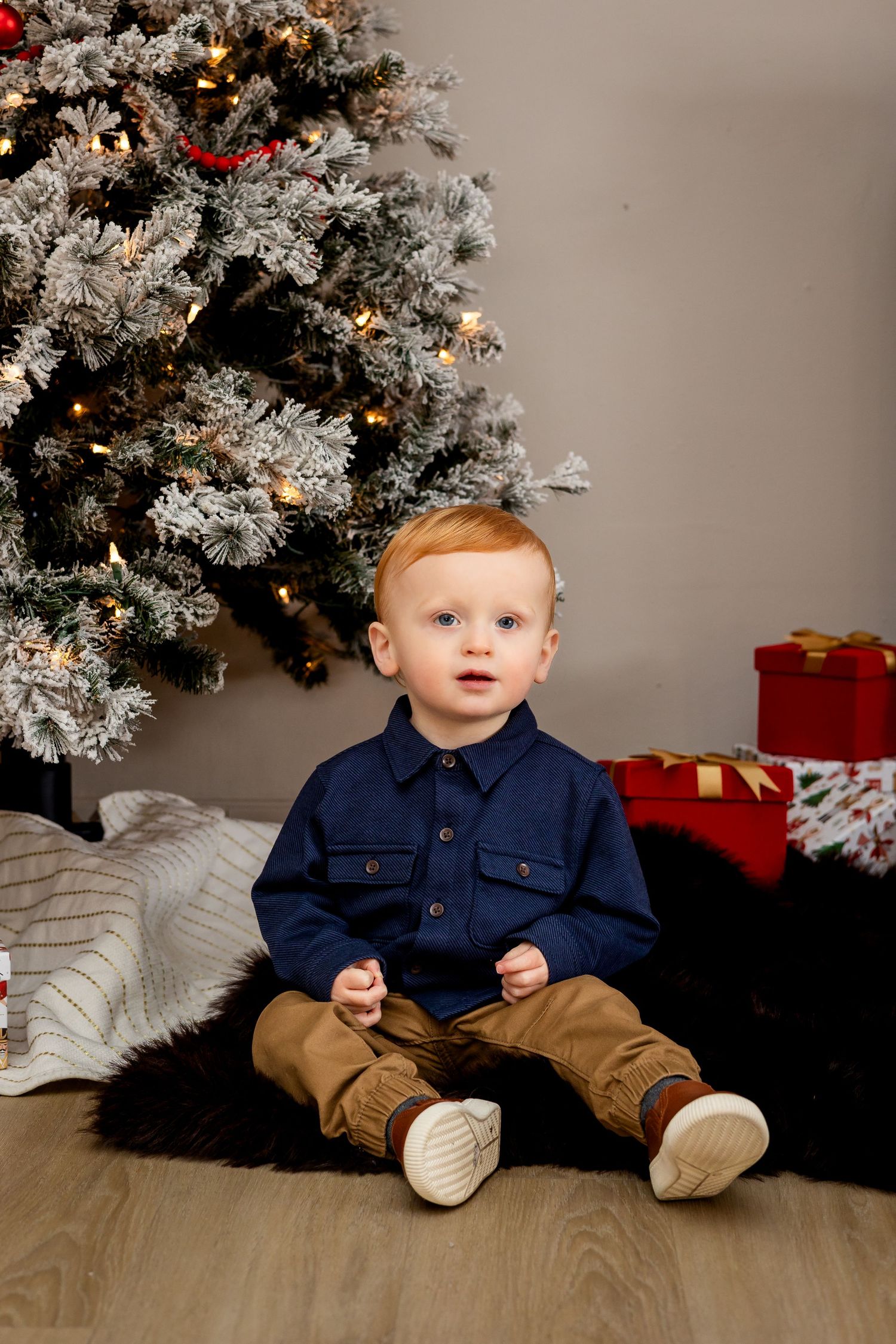 Cute toddler boy in navy shirt sits on fur rug by snow-flocked Christmas tree with gifts.