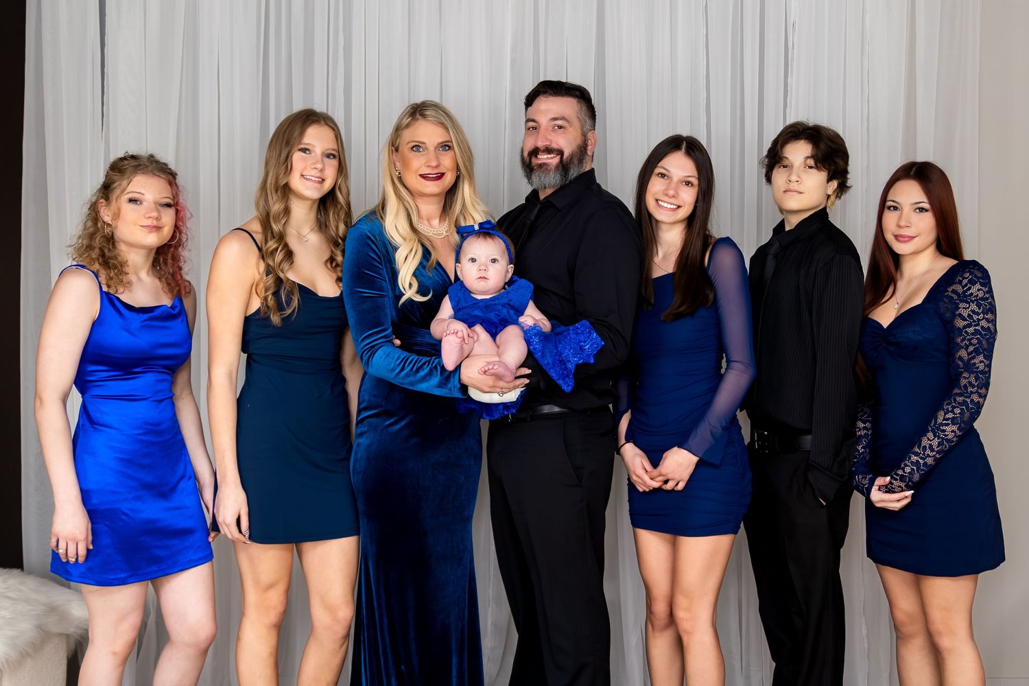 Family portrait in coordinating blue outfits, including a man holding a baby in blue, posing together indoors.