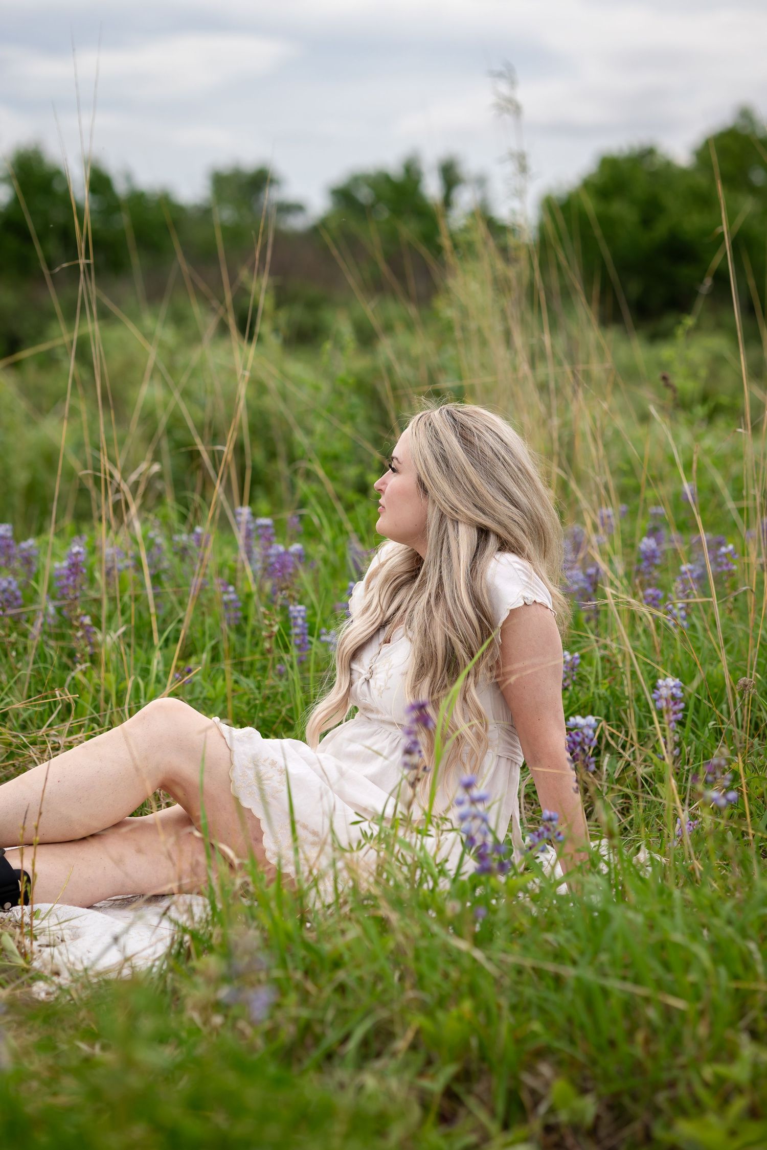 A young blonde woman in a white dress relaxes in a lush green field surrounded by purple wildflowers on a cloudy day.