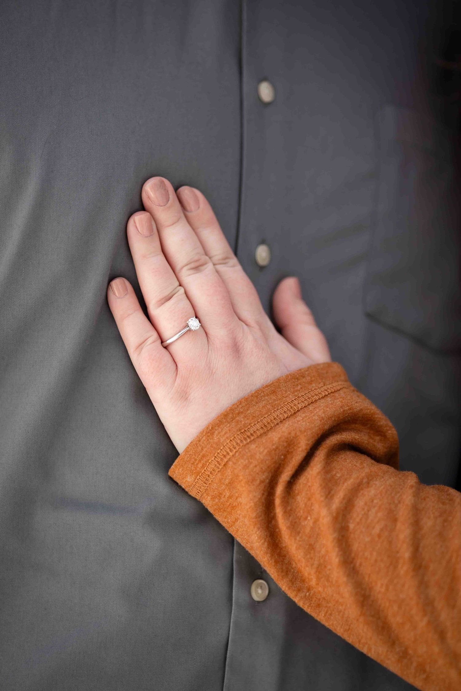 A hand wearing a diamond engagement ring rests on a man's grey button-up shirt, with an orange sweater sleeve visible.