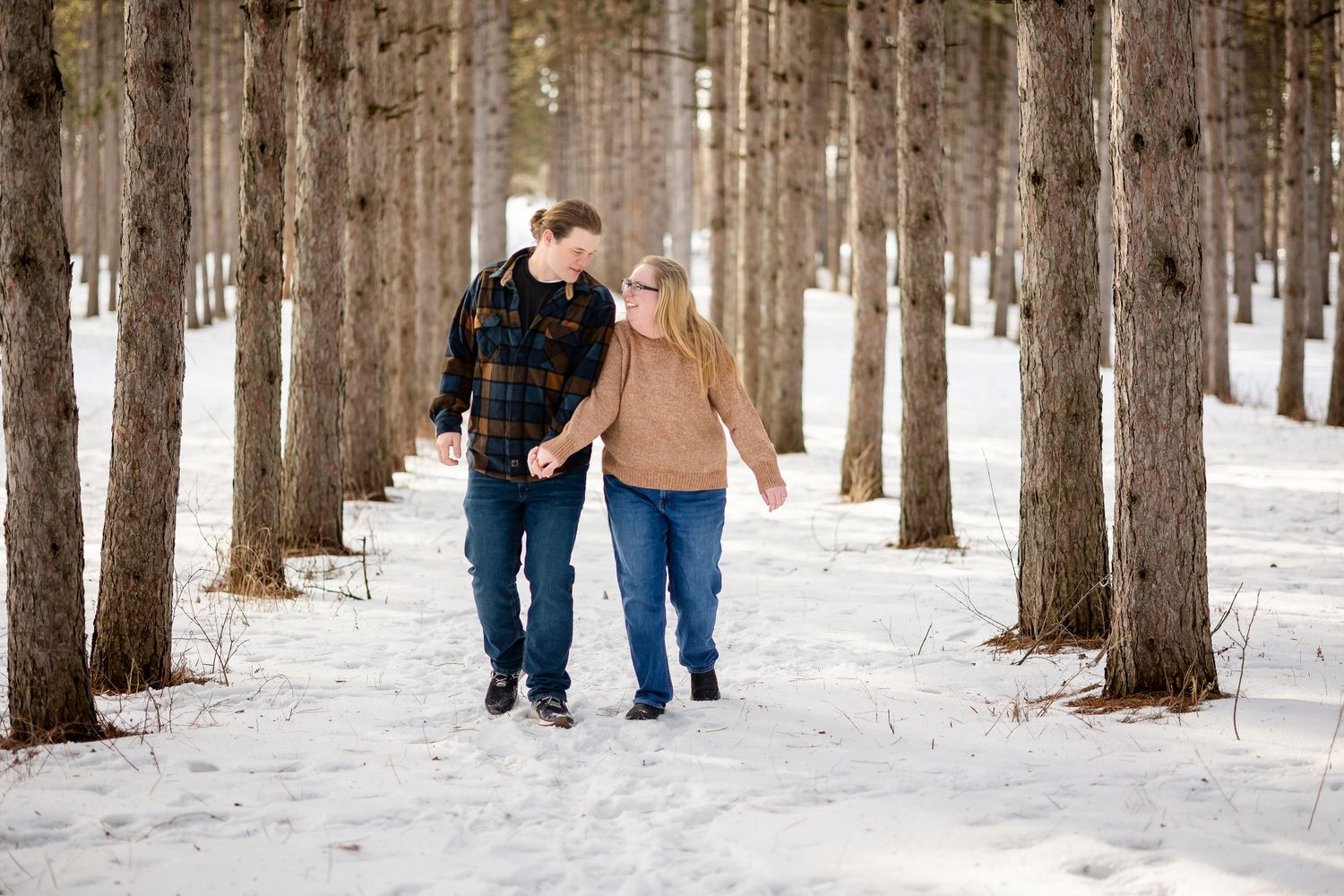Couple walking hand-in-hand through snowy pine forest in winter, wearing casual warm clothing.