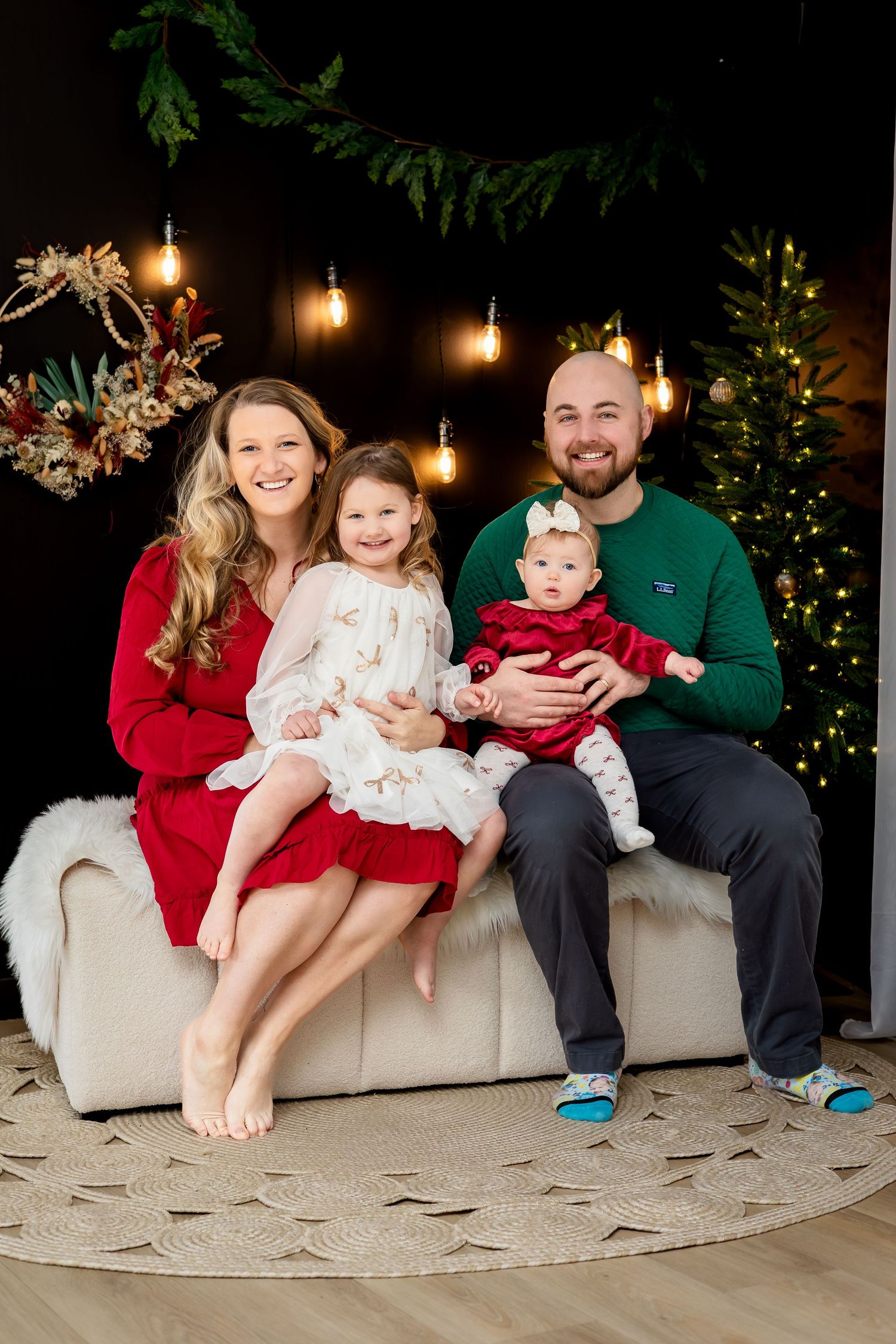 Family of four poses for Christmas portrait with holiday decorations, lights, and Christmas tree in background.