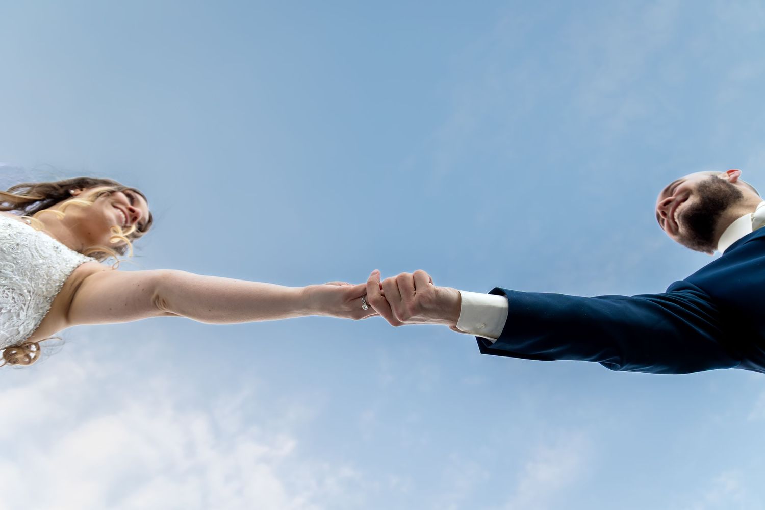 Bride and groom holding hands against blue sky, low angle wedding photo.
