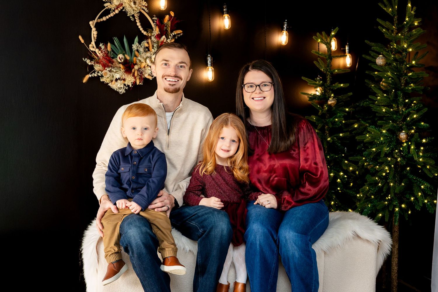 Family of four posing for a festive Christmas portrait with holiday lights and decorated tree in background.