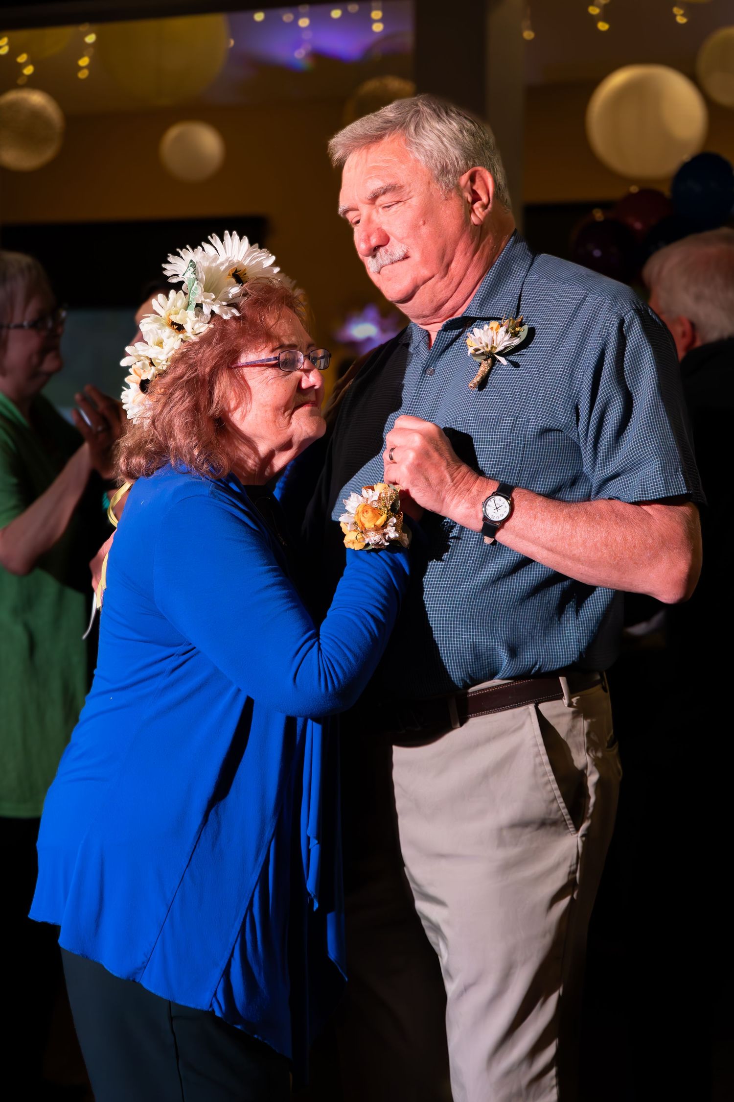 A couple shares a dance moment under party lights, wearing blue and khaki with floral accessories.
